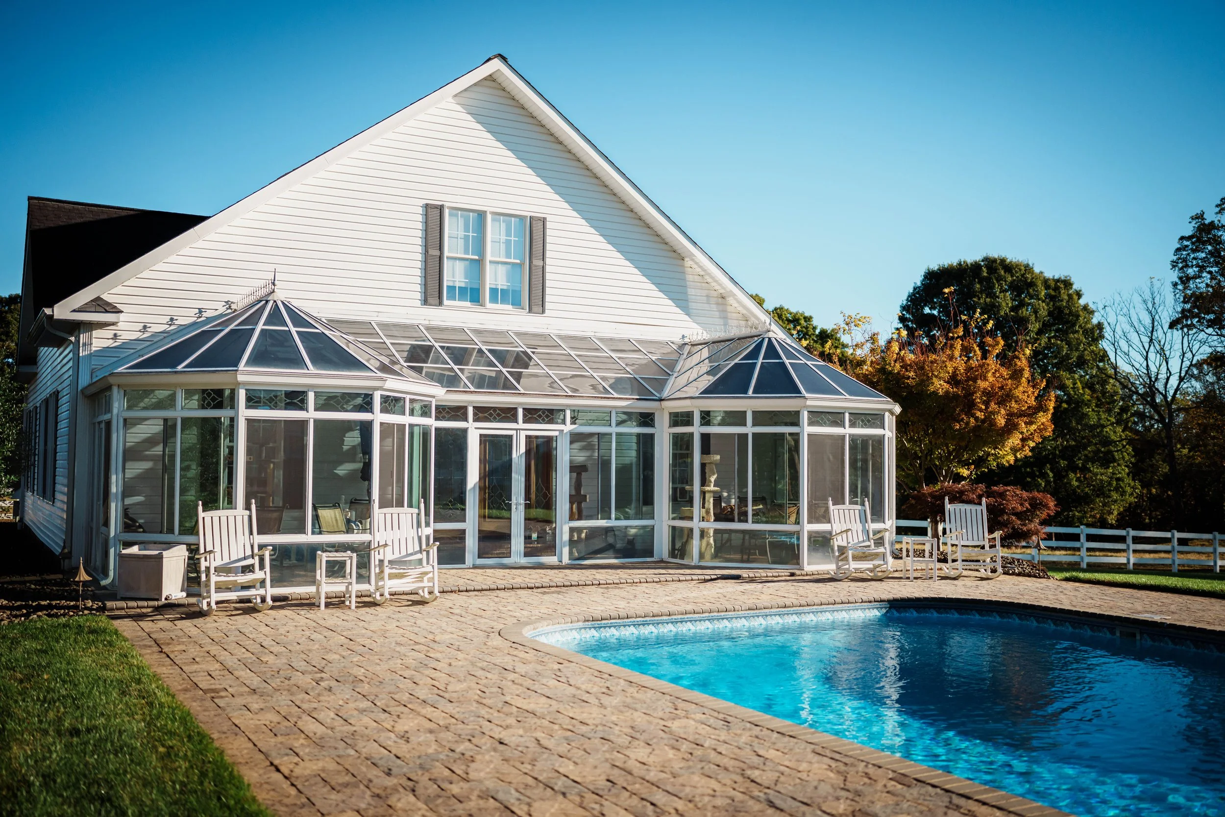 White house with a glass conservatory attached, brick patio with outdoor chairs, and a swimming pool, surrounded by trees and a white fence under a clear blue sky.