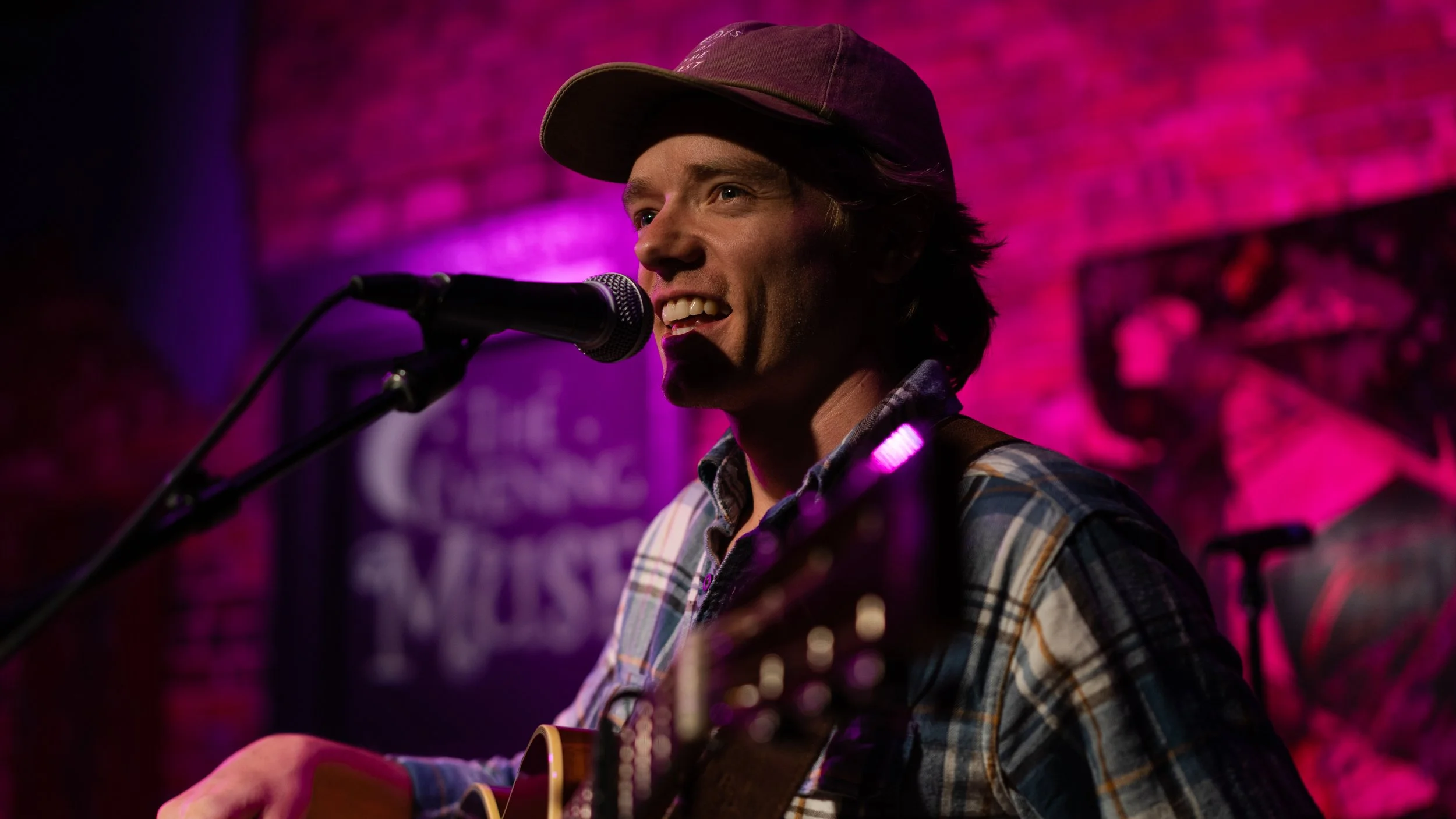 A male musician in a plaid shirt and baseball cap singing into a microphone while playing an acoustic guitar on a stage with pink lighting and a brick wall background.