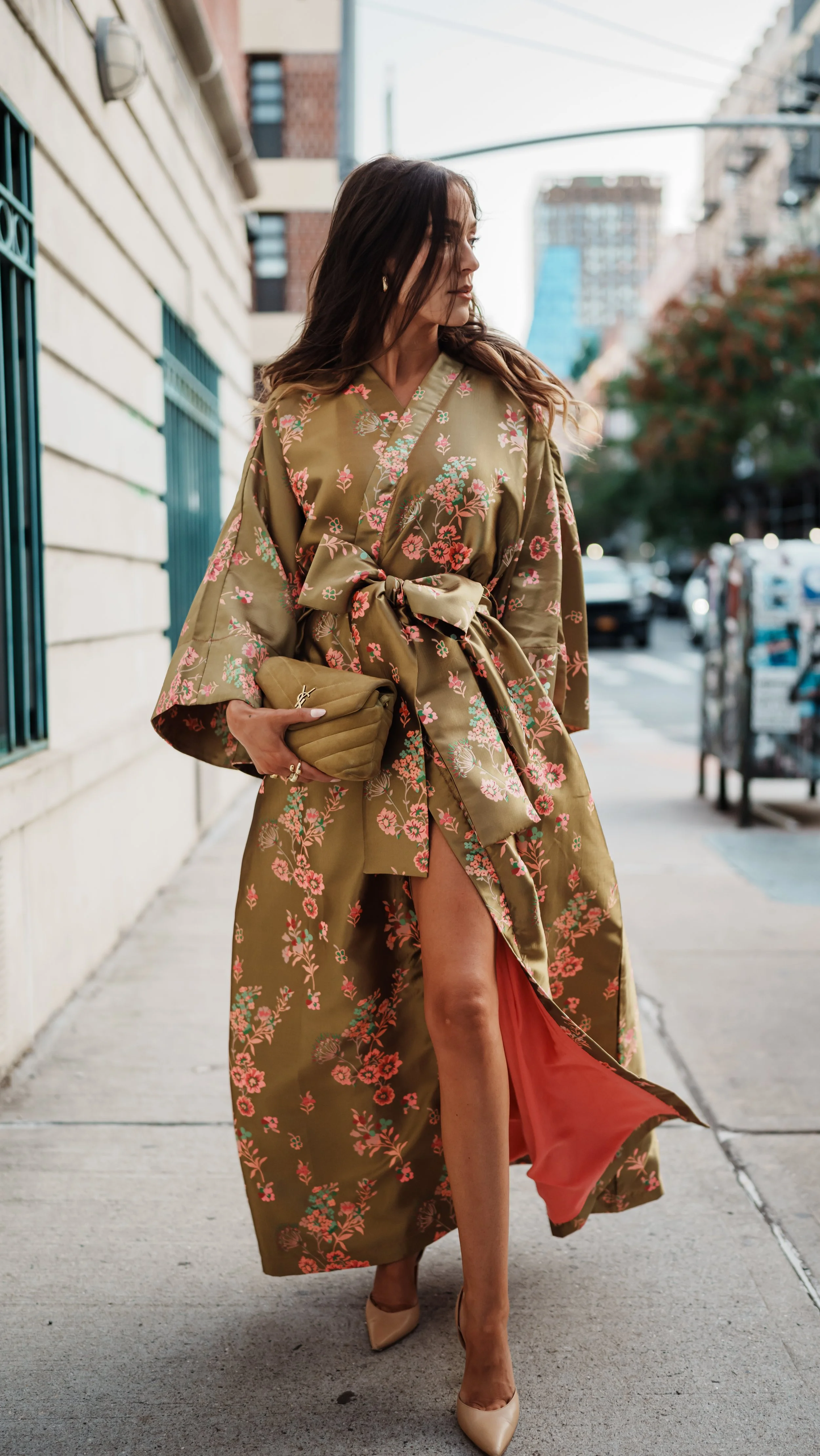 A woman walking on a city sidewalk wearing a long, satin floral kimono with a sash tie, beige high heels, and holding a matching clutch purse.