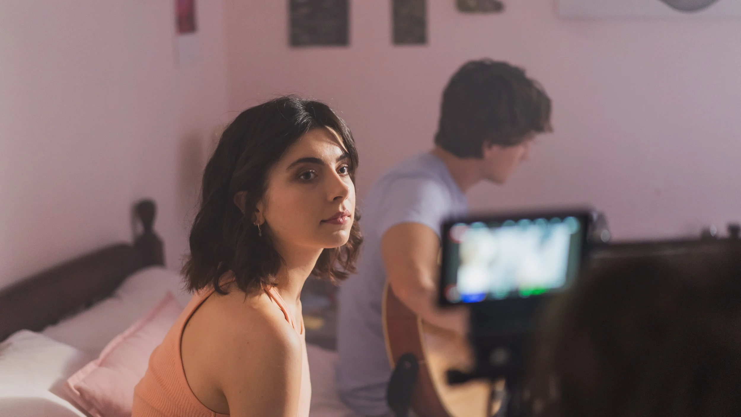 A young woman with shoulder-length dark hair and earrings sitting on bed, looking at the camera, with a young man playing guitar in the background, a camera recording them in a softly lit room.
