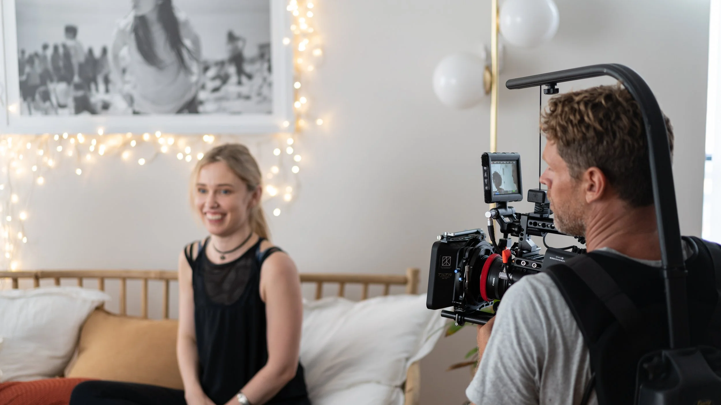 A woman in black clothing smiling for a camera while sitting on a bed, with a man operating professional filming equipment capturing her in a well-lit room decorated with string lights and a large black and white photograph.