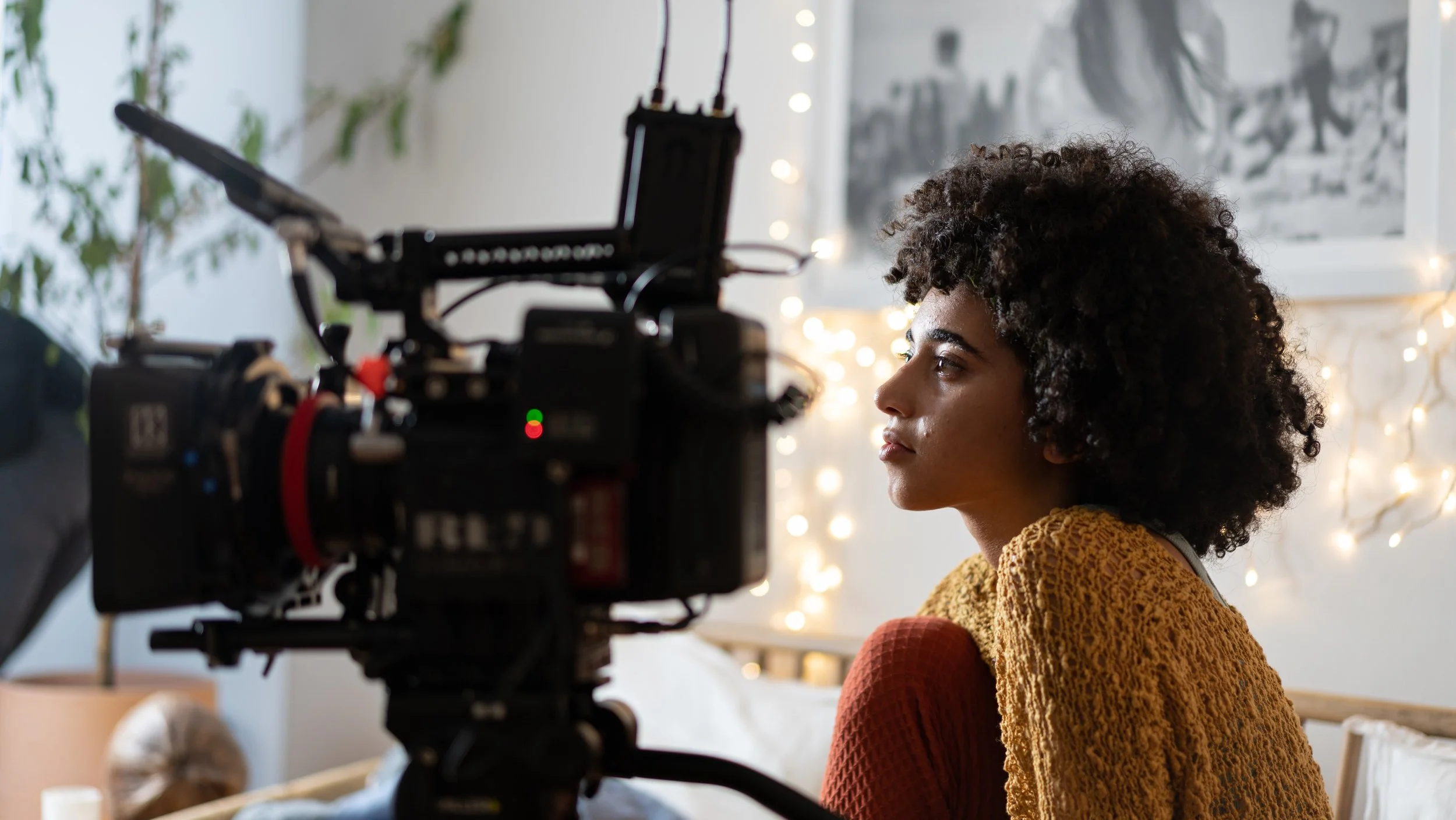 A woman with curly dark hair sits in front of a camera, with fairy lights and artwork in the background.
