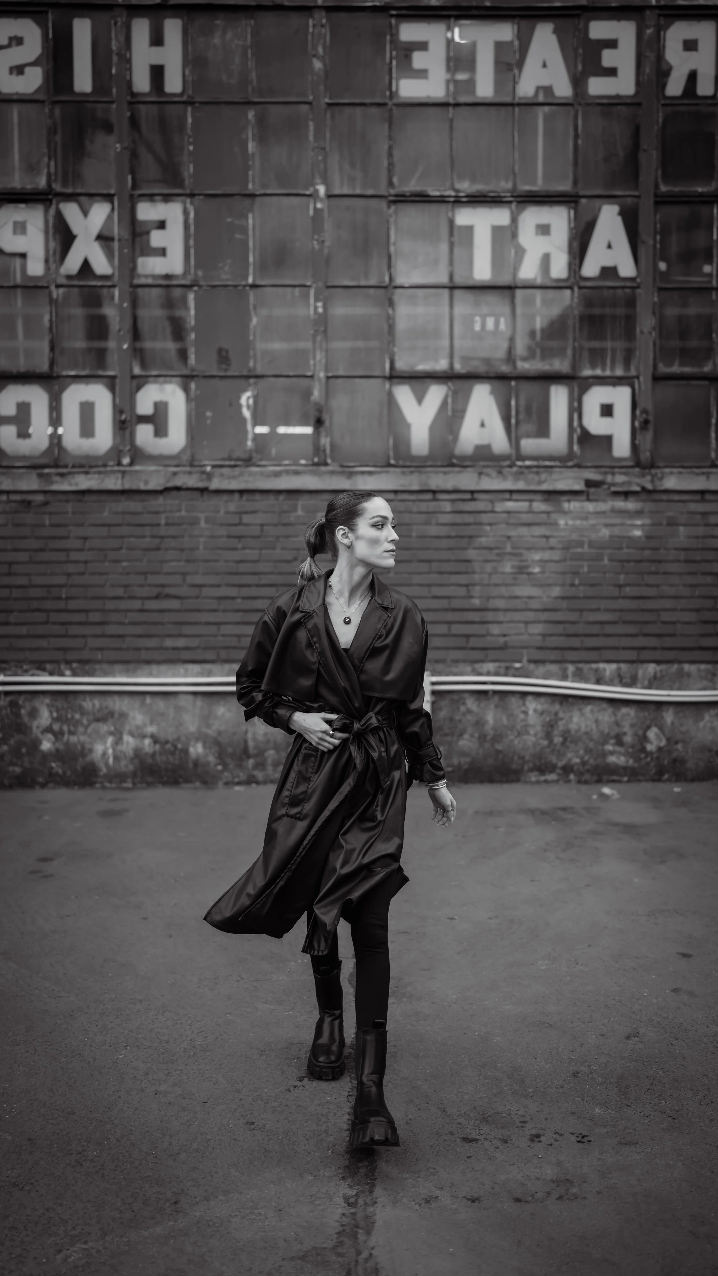 A woman walking outdoors in an urban setting with old brick wall and large window in the background. The scene is in grayscale.