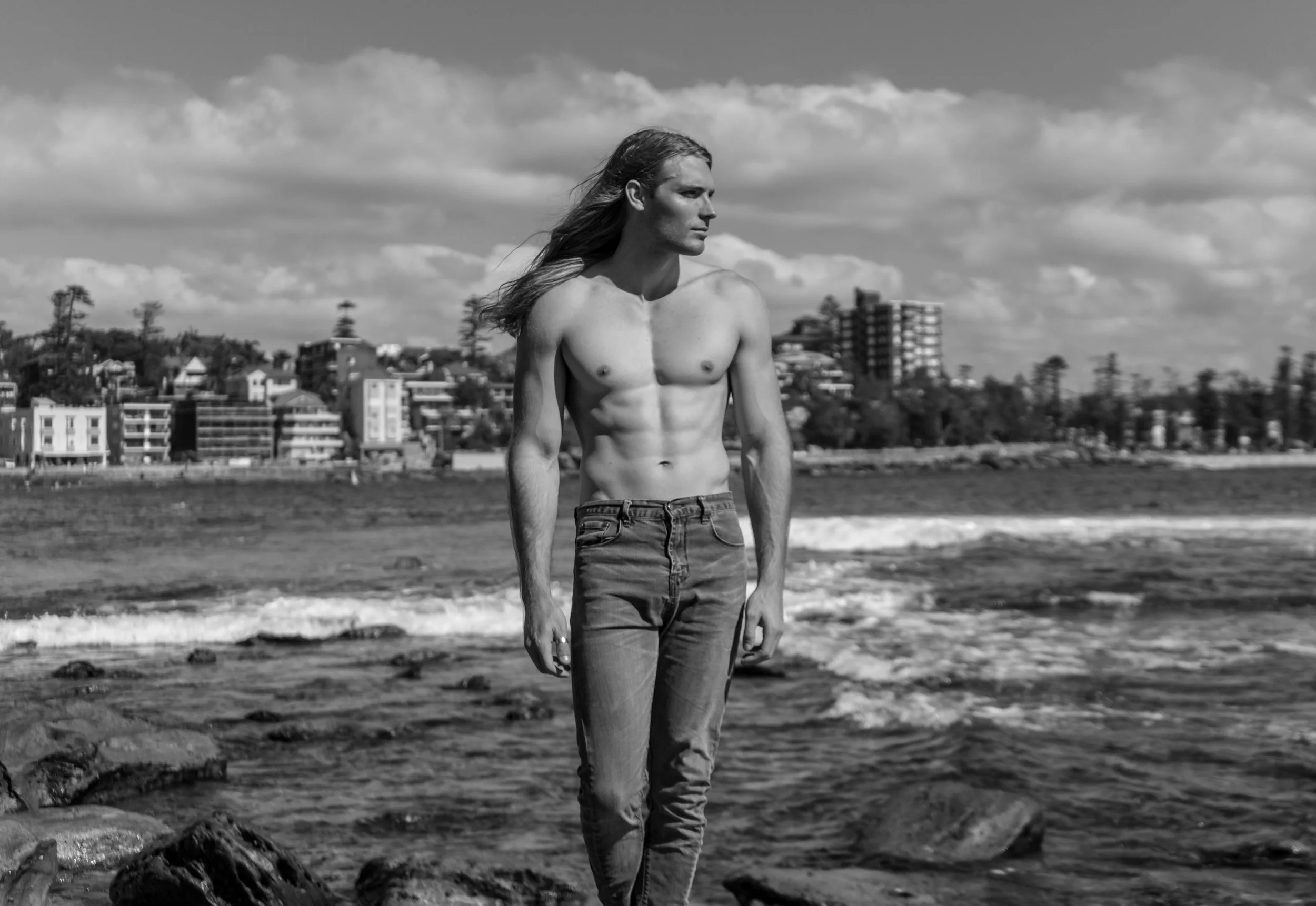 A shirtless man with long hair standing on rocks at the beach with waves in the background, black and white photo.