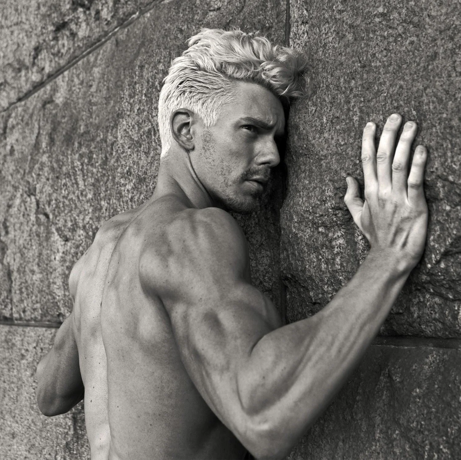 Black and white photo of a muscular man with bleached blonde hair, shirtless, pressing his side against a rough stone wall, with his right hand touching the wall.