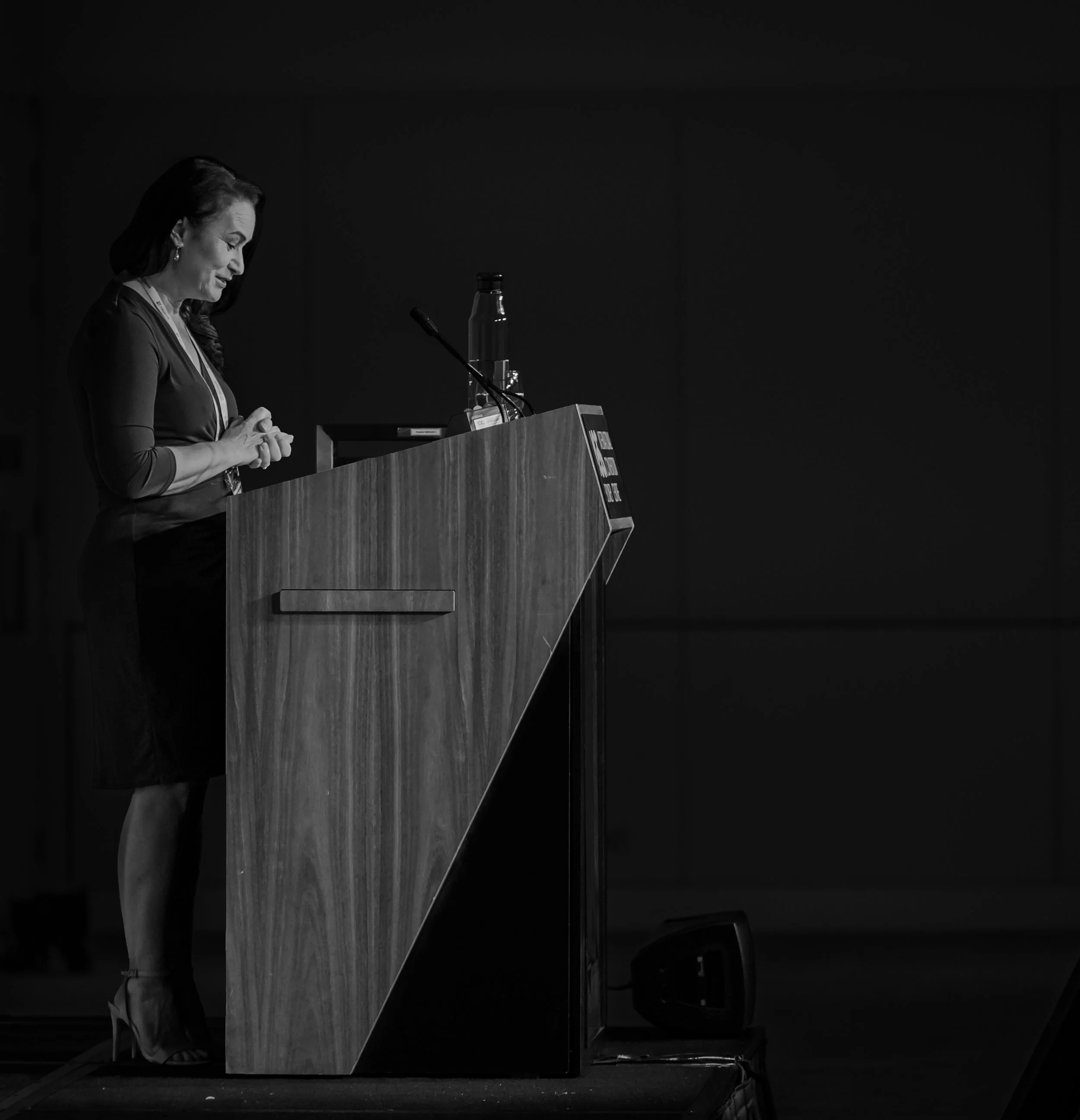 A woman stands at a wooden podium on a stage, looking down with her hands clasped, in a dimly lit room.