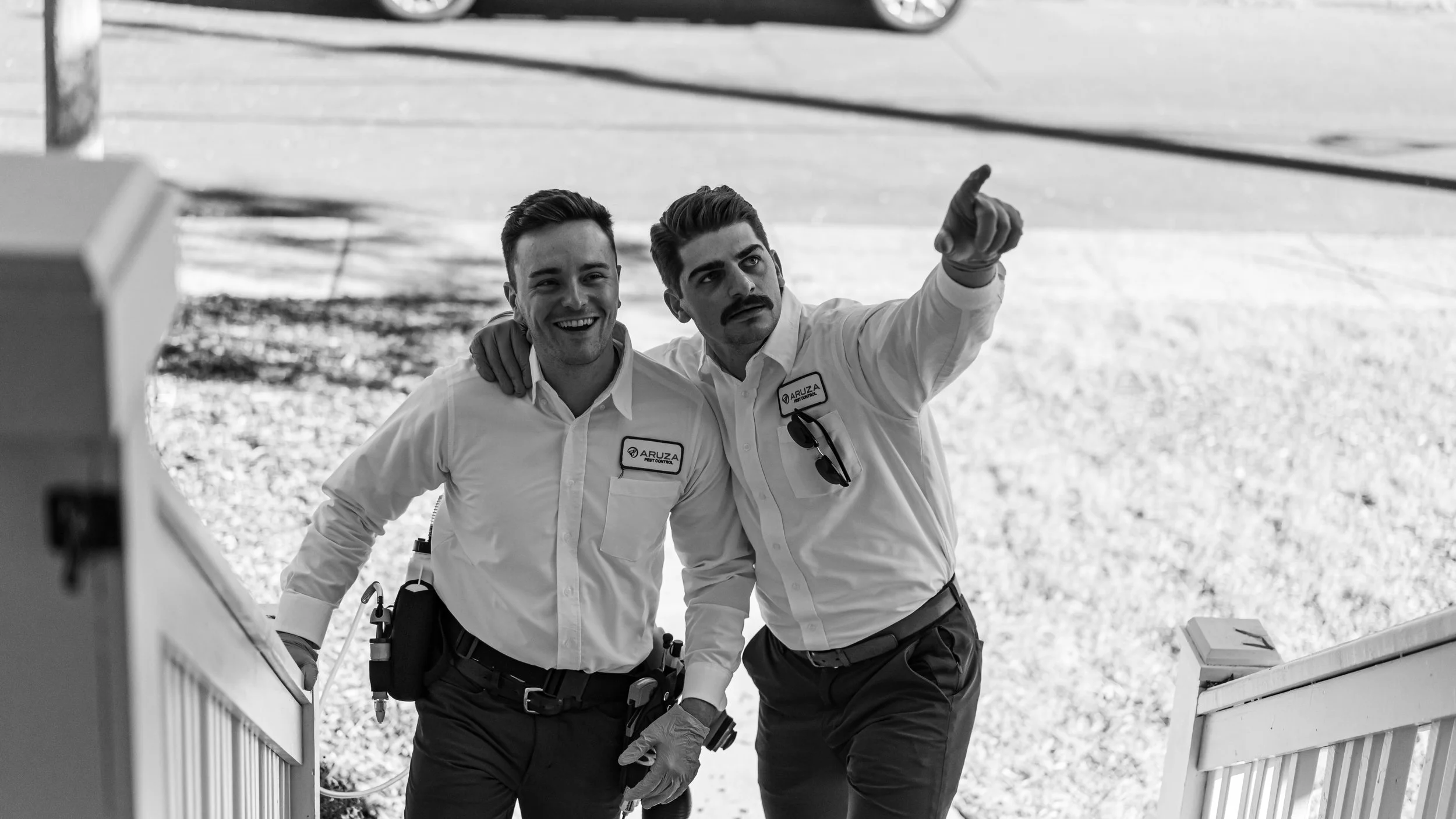 Two male workers, one with a moustache, smiling and pointing, standing outside near a staircase railing, wearing white shirts with company logos and tools attached, in black and white.