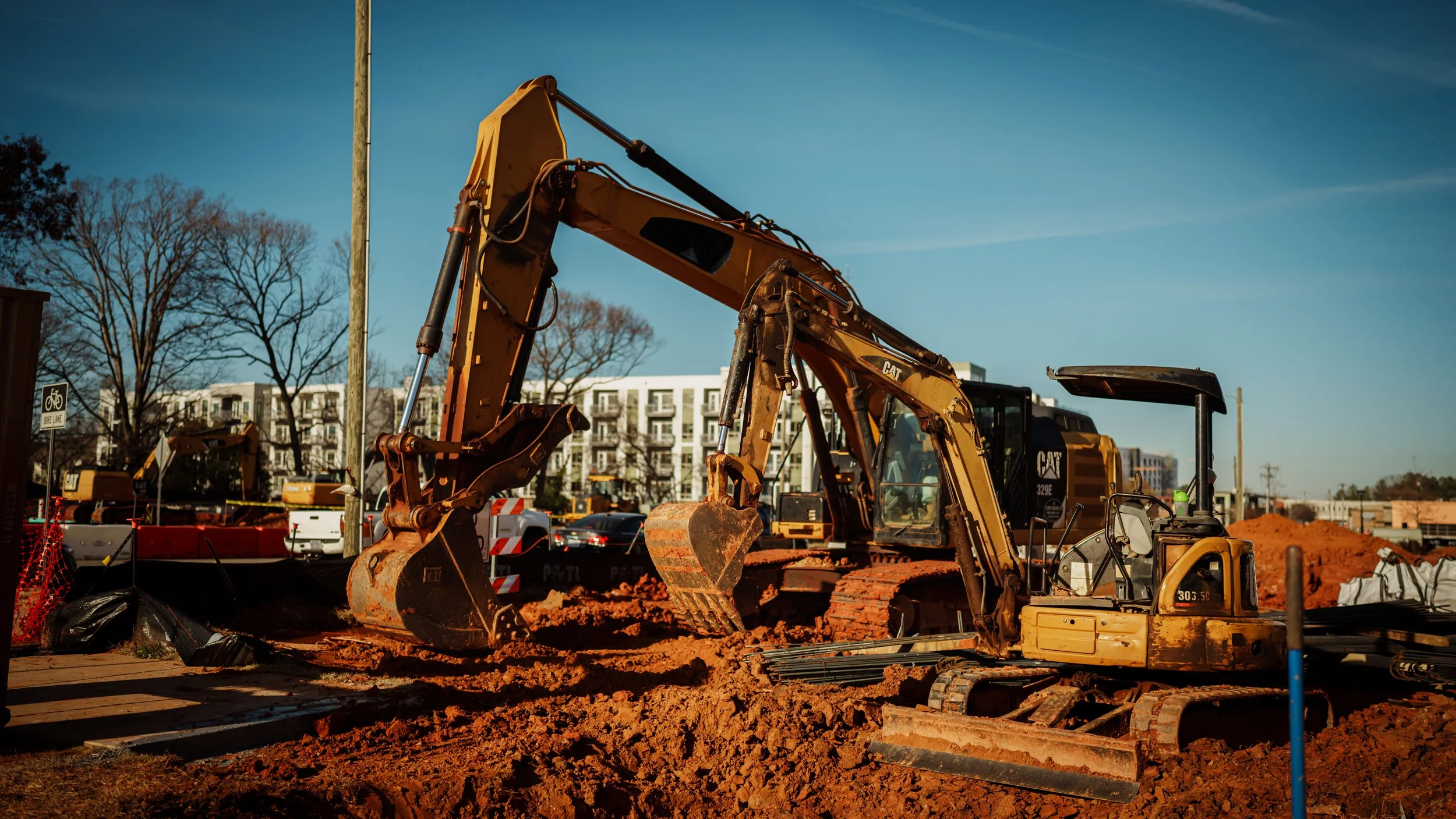 A yellow excavator at a construction site digging into reddish-orange soil with construction barriers and equipment around, and trees and buildings in the background.