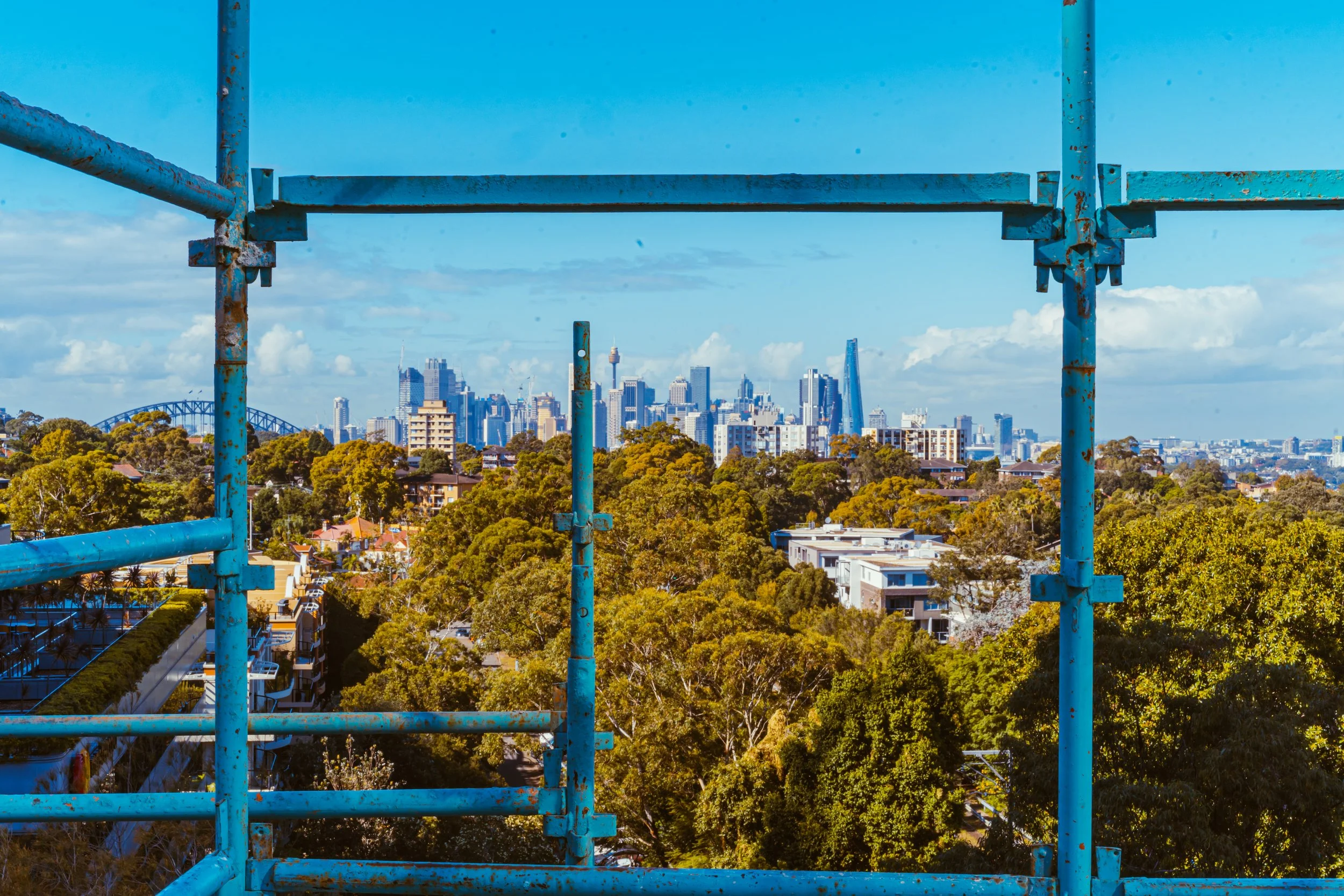 View of a city skyline through a blue, rusted scaffolding with green trees in the foreground and a bright blue sky.