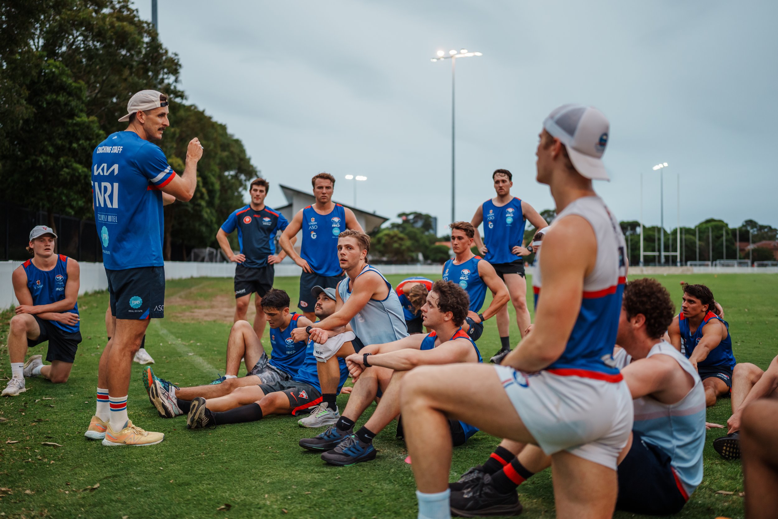 A group of young male athletes in sports uniforms sitting and kneeling on a grassy field, listening to a coach during a team practice or meeting.