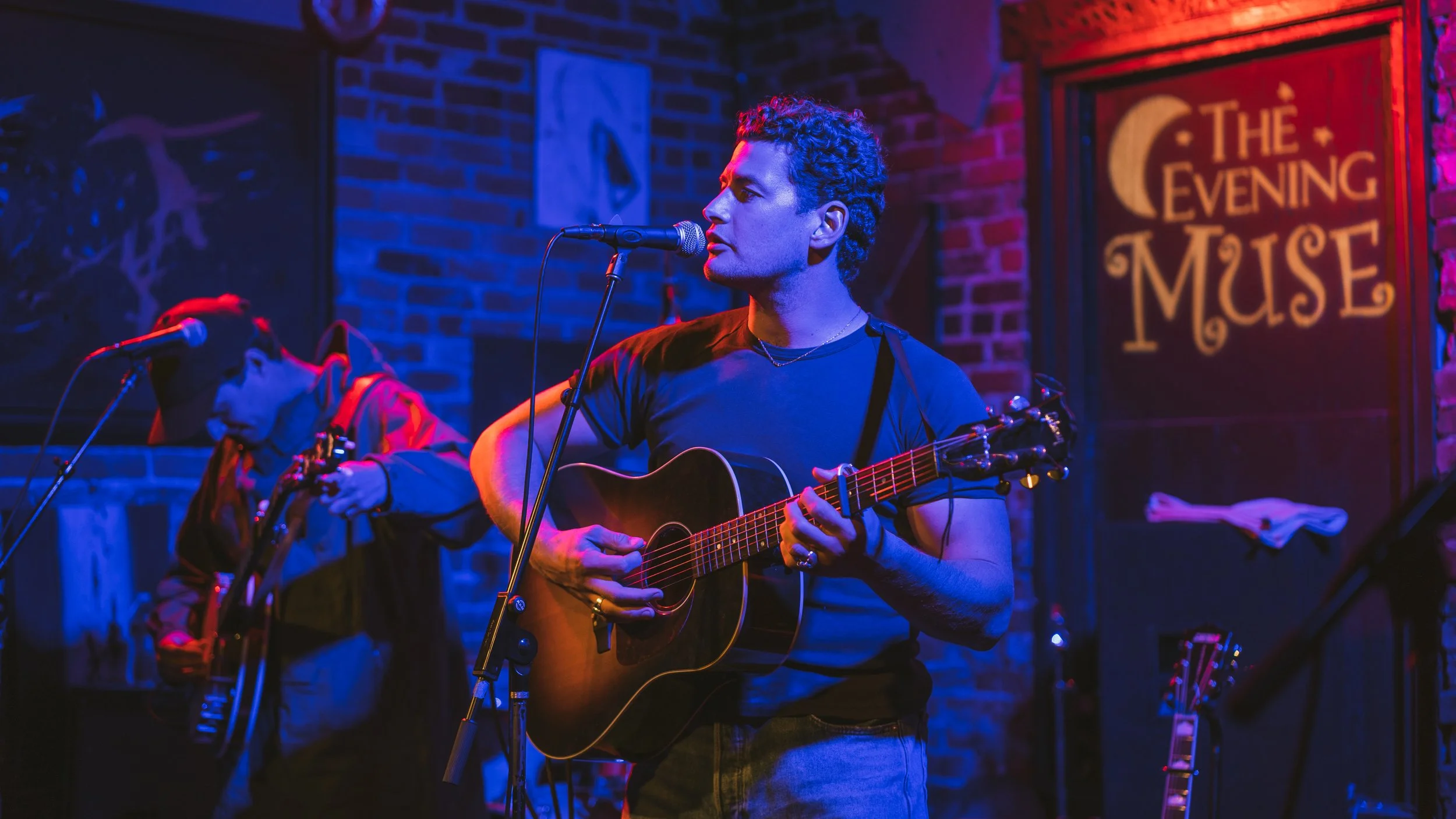 A man playing an acoustic guitar and singing at a live music venue called The Evening Muse, with brick walls and artwork in the background.