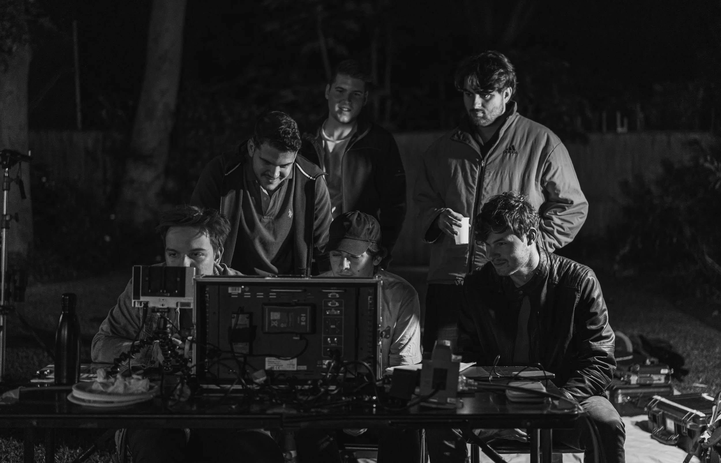 Group of six young men working on a film or video production setup outdoors at night, gathered around a monitor and various equipment.