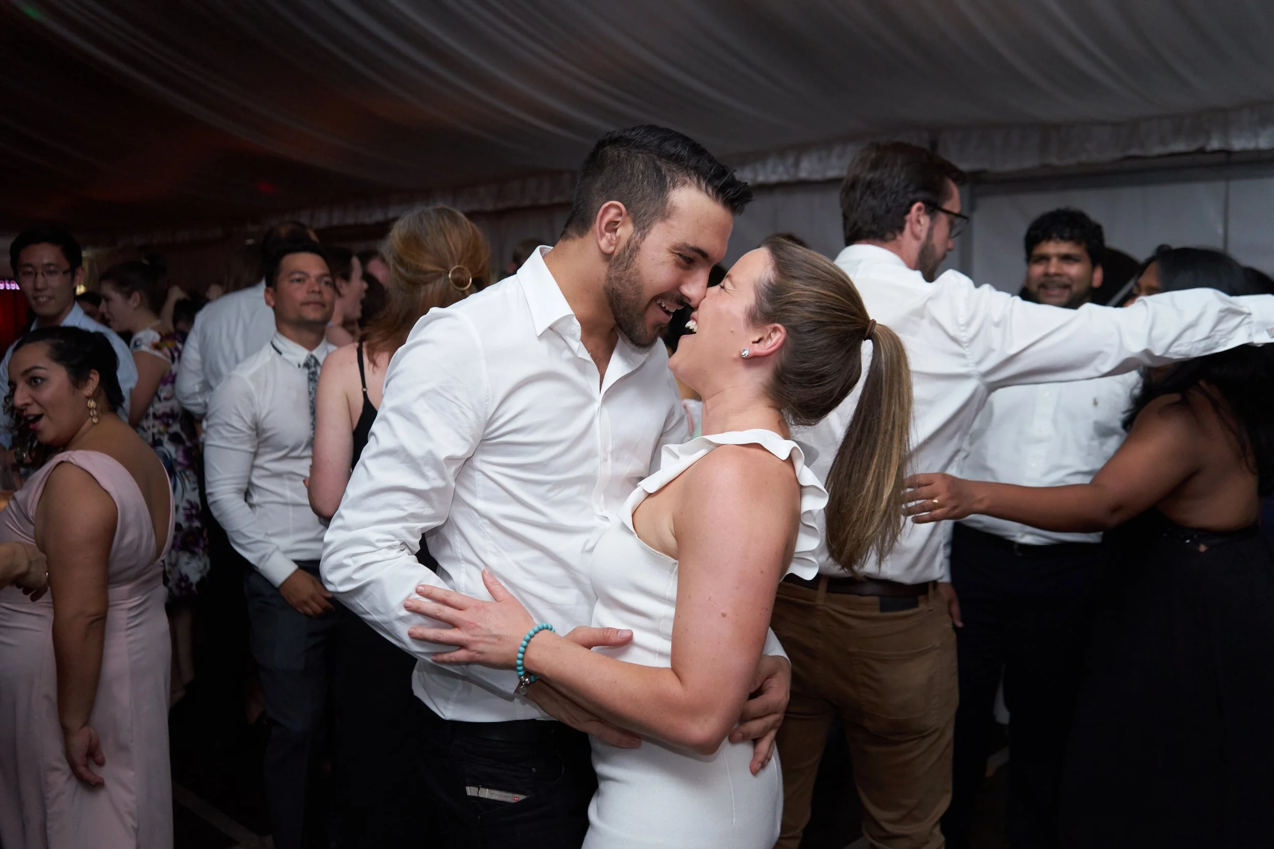 A couple dancing closely, smiling and touching foreheads at a festive event with other people dancing in the background.