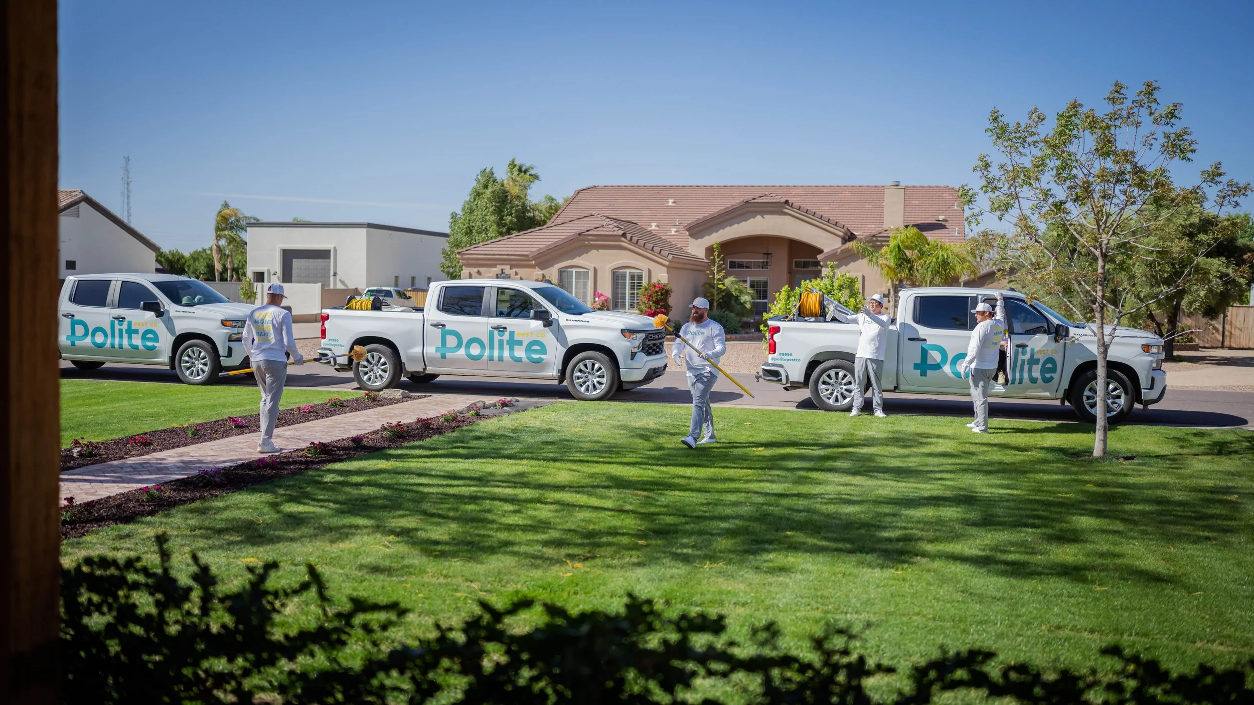 Four workers in white uniforms spraying pesticide in front of a residential house, with three white trucks labeled 'Polite' parked nearby on the street.