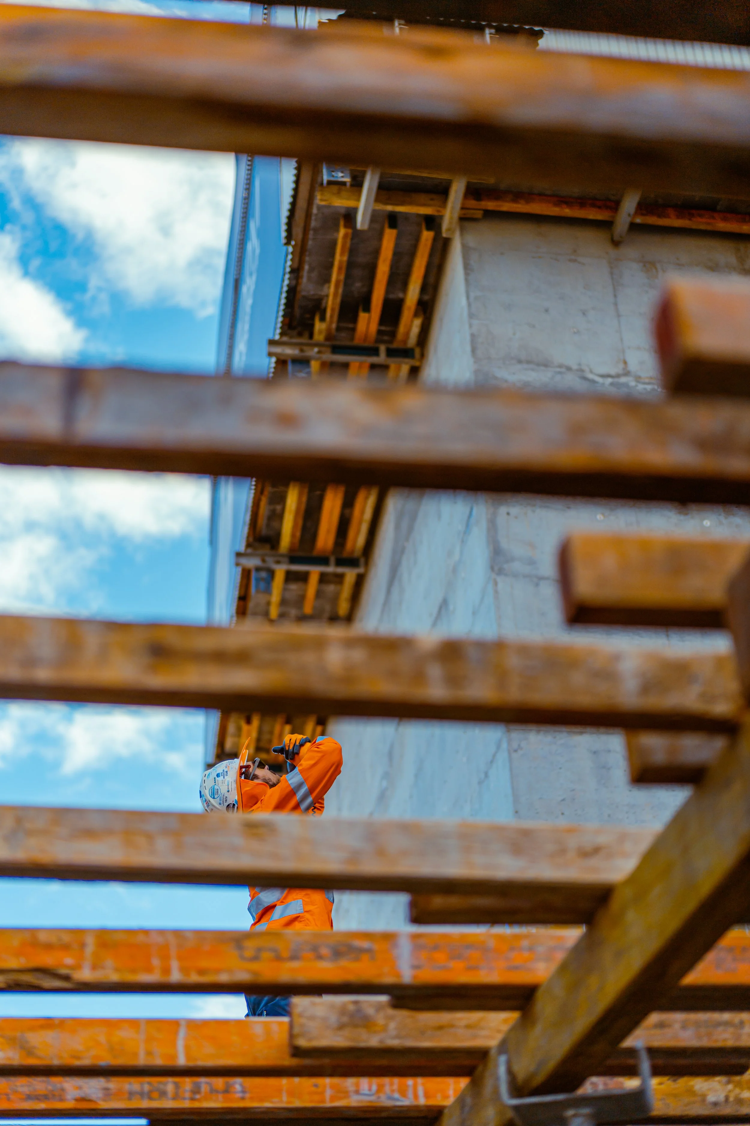 Construction worker in orange safety gear and helmet taking a photo or inspecting the construction site from beneath wooden beams.