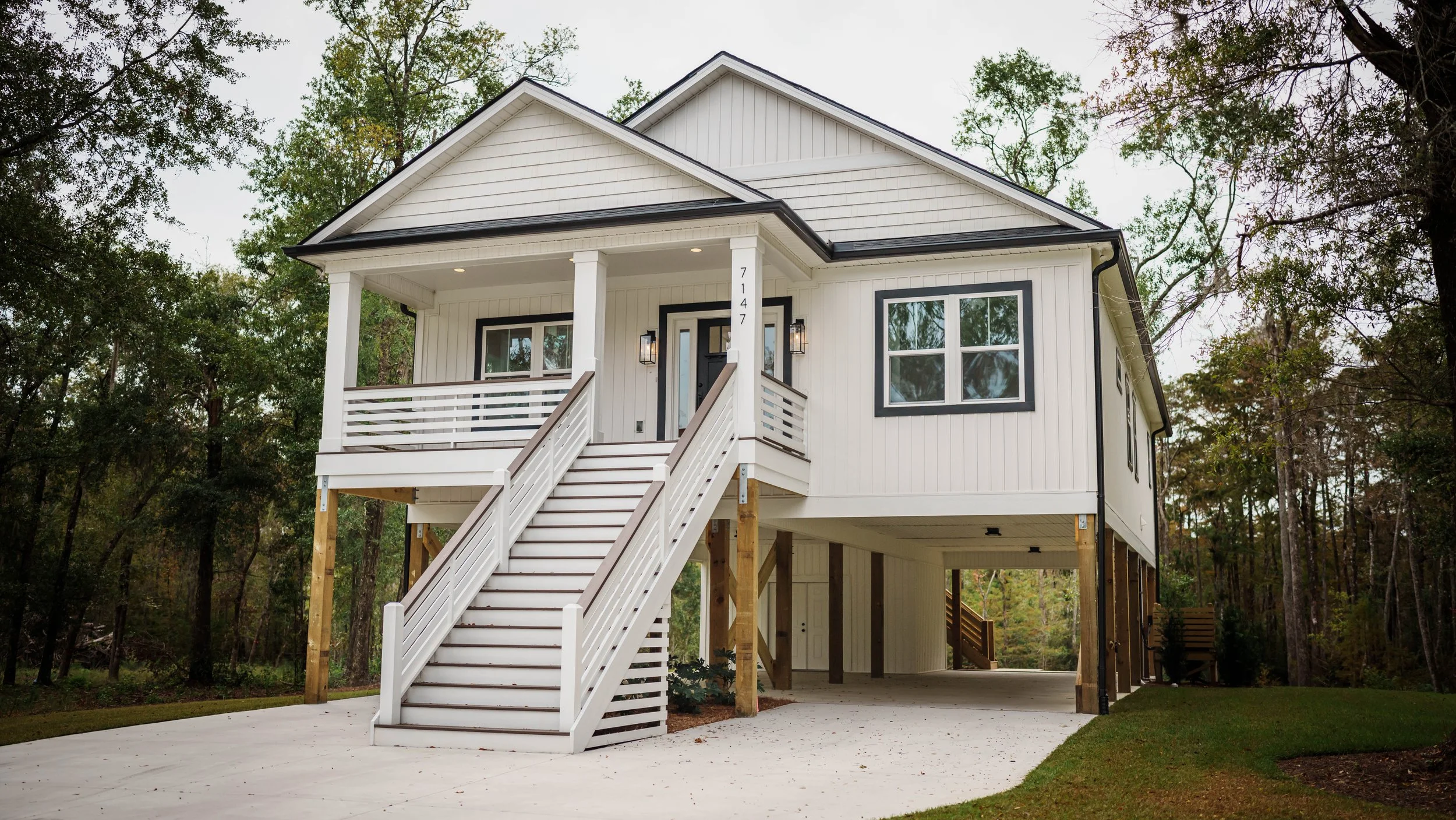 A white raised house with a front porch, stairs, and black trim, surrounded by trees.