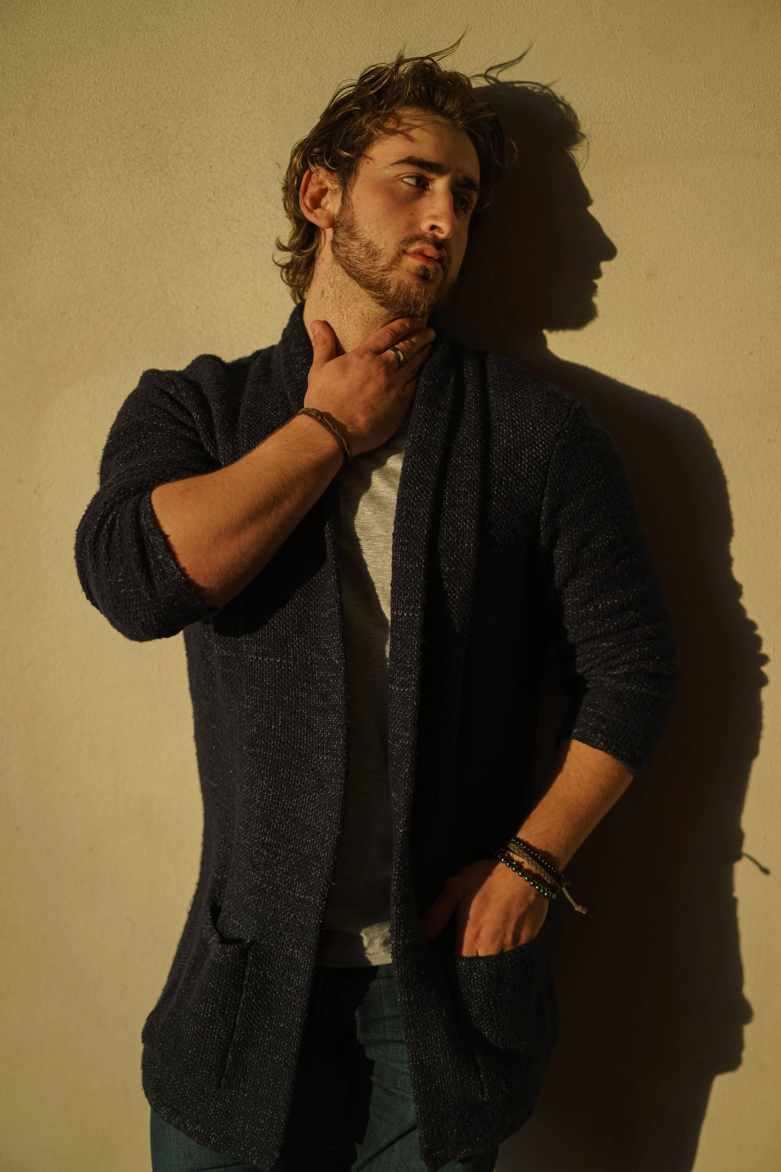 A young man with wavy hair and a beard standing against a beige wall, casting a shadow to his right. He wears a dark textured blazer over a light shirt, with bracelets on his left wrist. He touches his neck with his right hand and looks to the left with a contemplative expression.