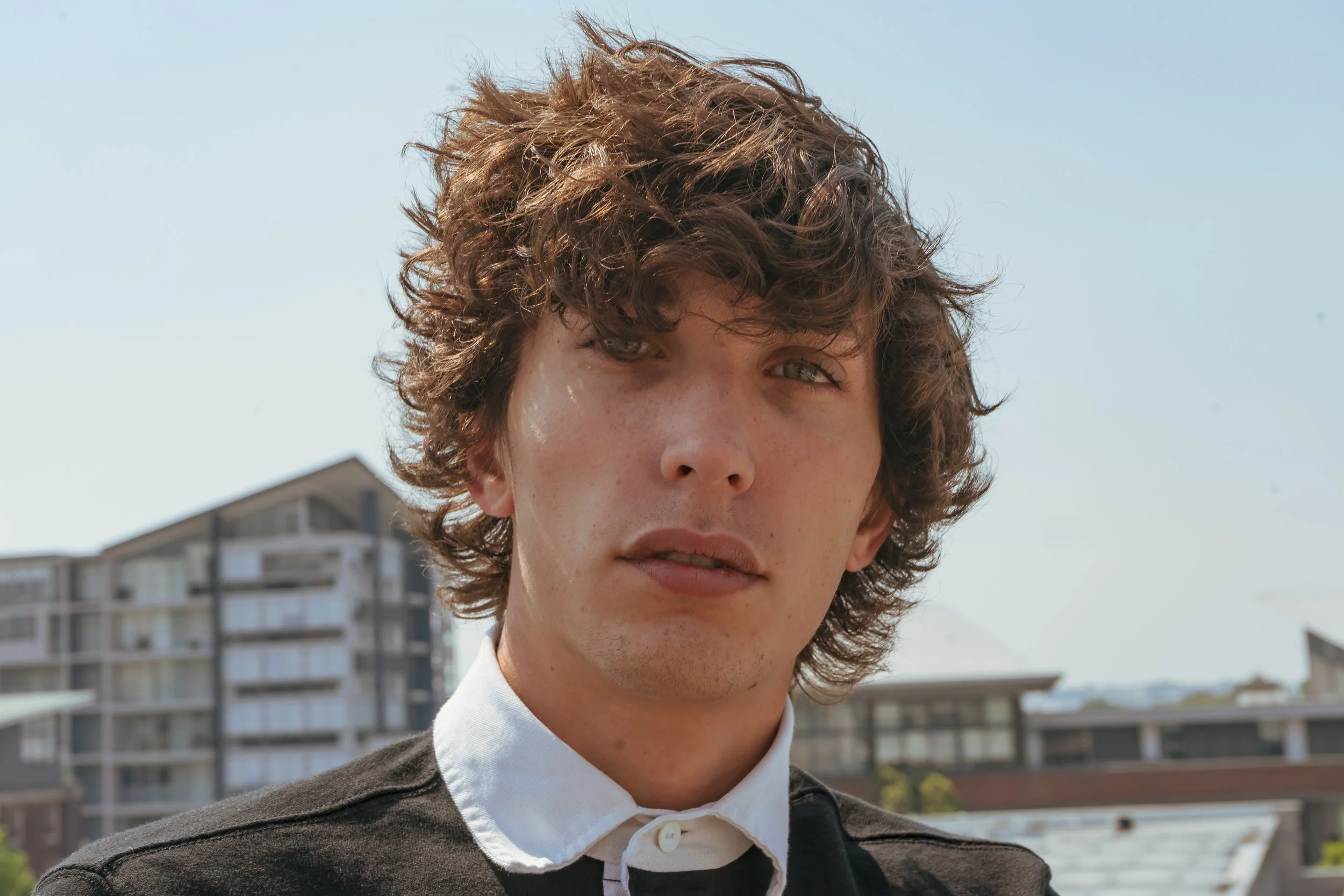 Close-up of a young man with curly brown hair and light skin, wearing a black top with a white collar, outdoors with buildings and clear sky in the background.