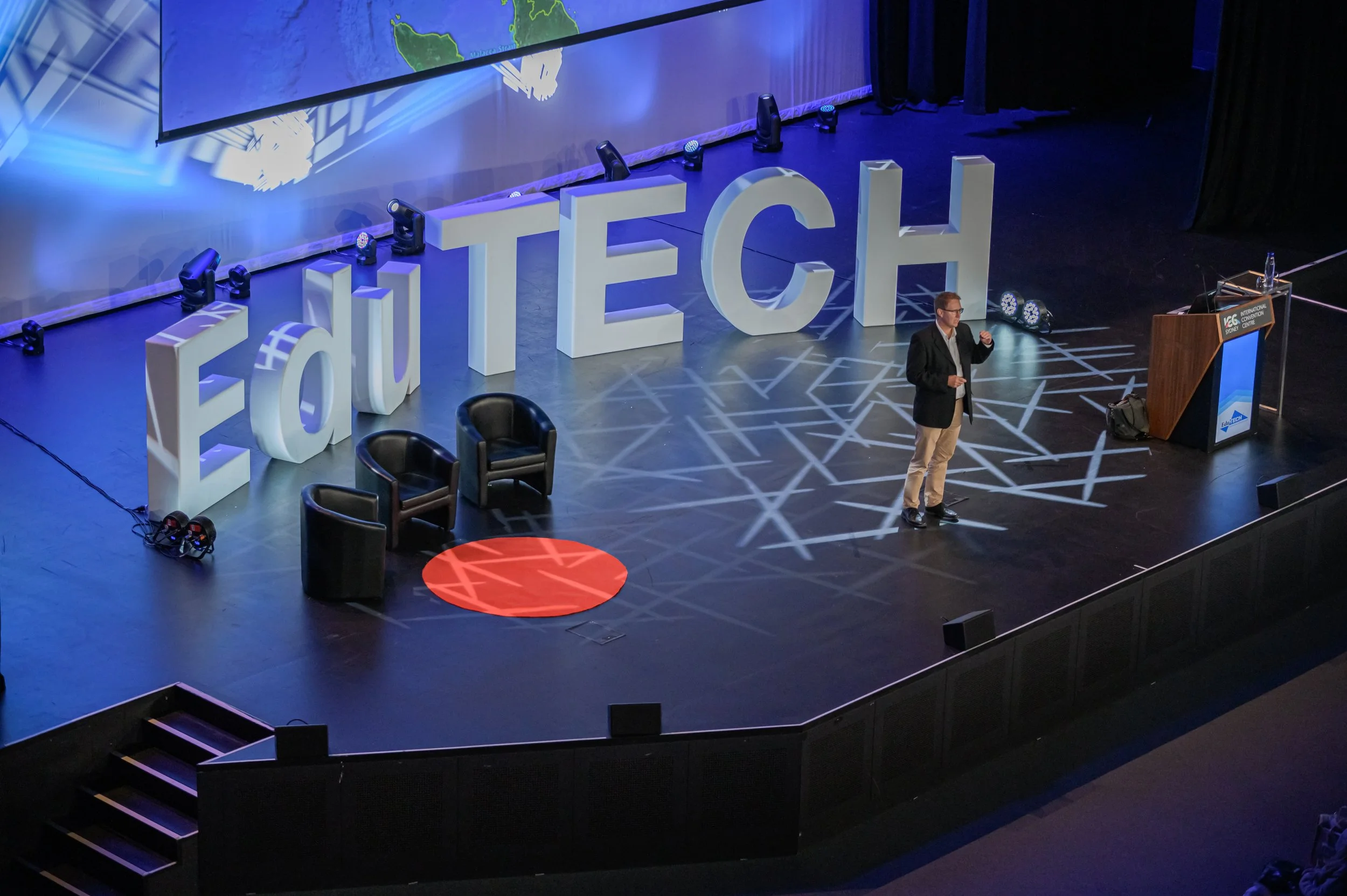 Stage setup with large 3D letters spelling 'EduTECH', a speaker standing on the stage, and seating arrangements with three chairs. A large screen displays a map in the background.