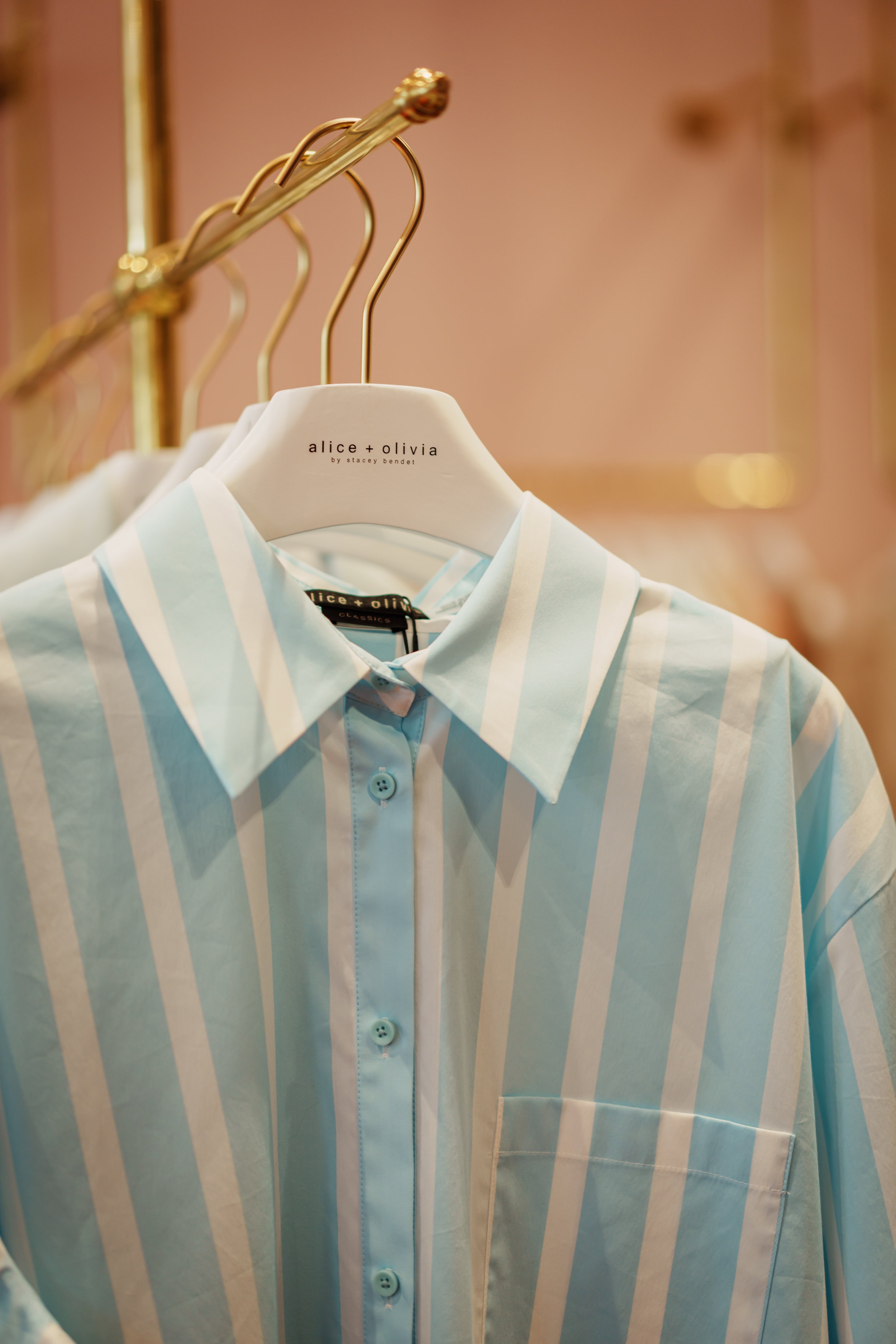 A light blue and white striped button-up shirt hanging on a white hanger in a clothing store. The hanger has the brand name 'alice + olivia' printed on it.