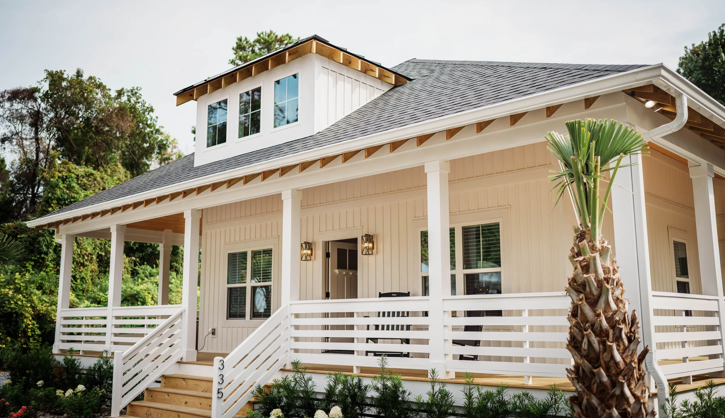 A white modern house with a front porch, stairs, and a palm tree in the foreground, surrounded by greenery.