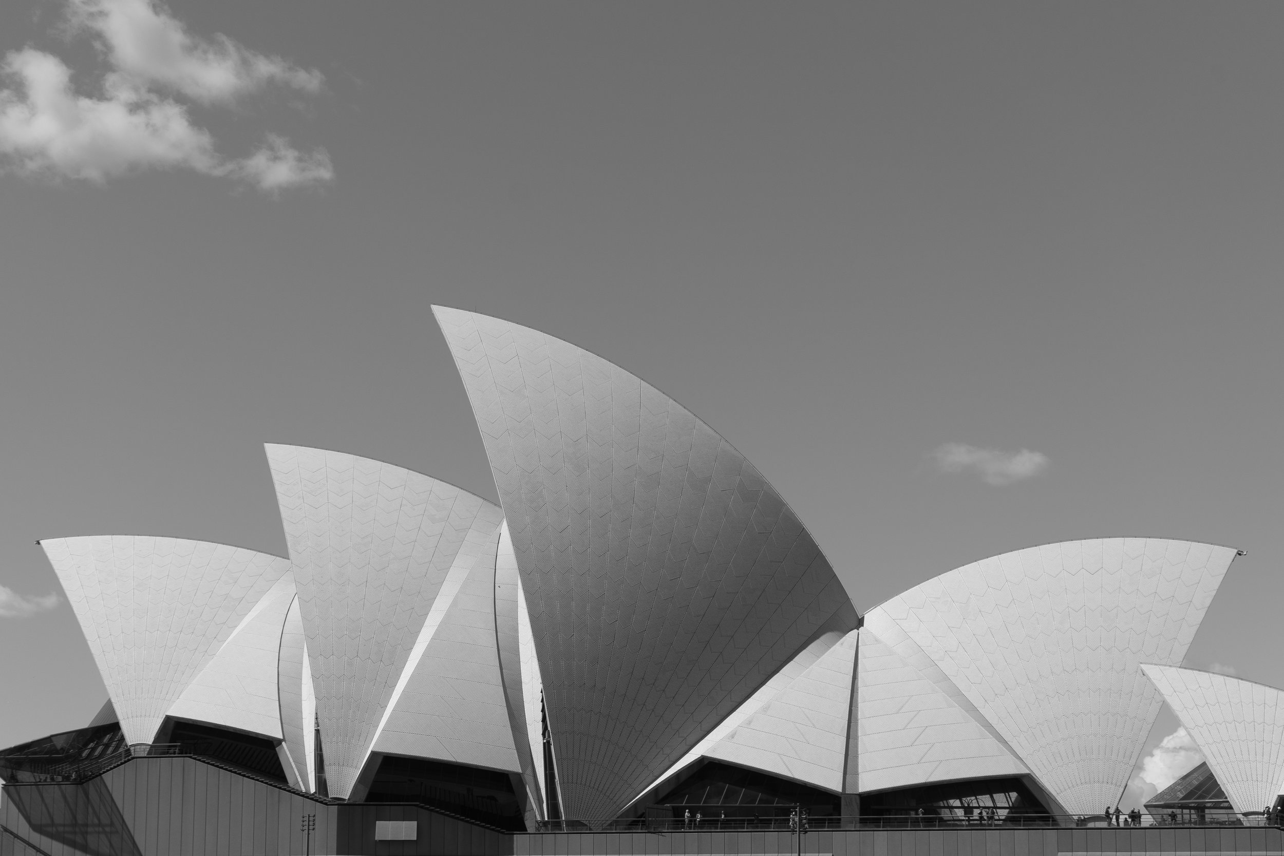 Black and white photo of the Sydney Opera House with its distinctive sail-like roof structures under a partly cloudy sky.