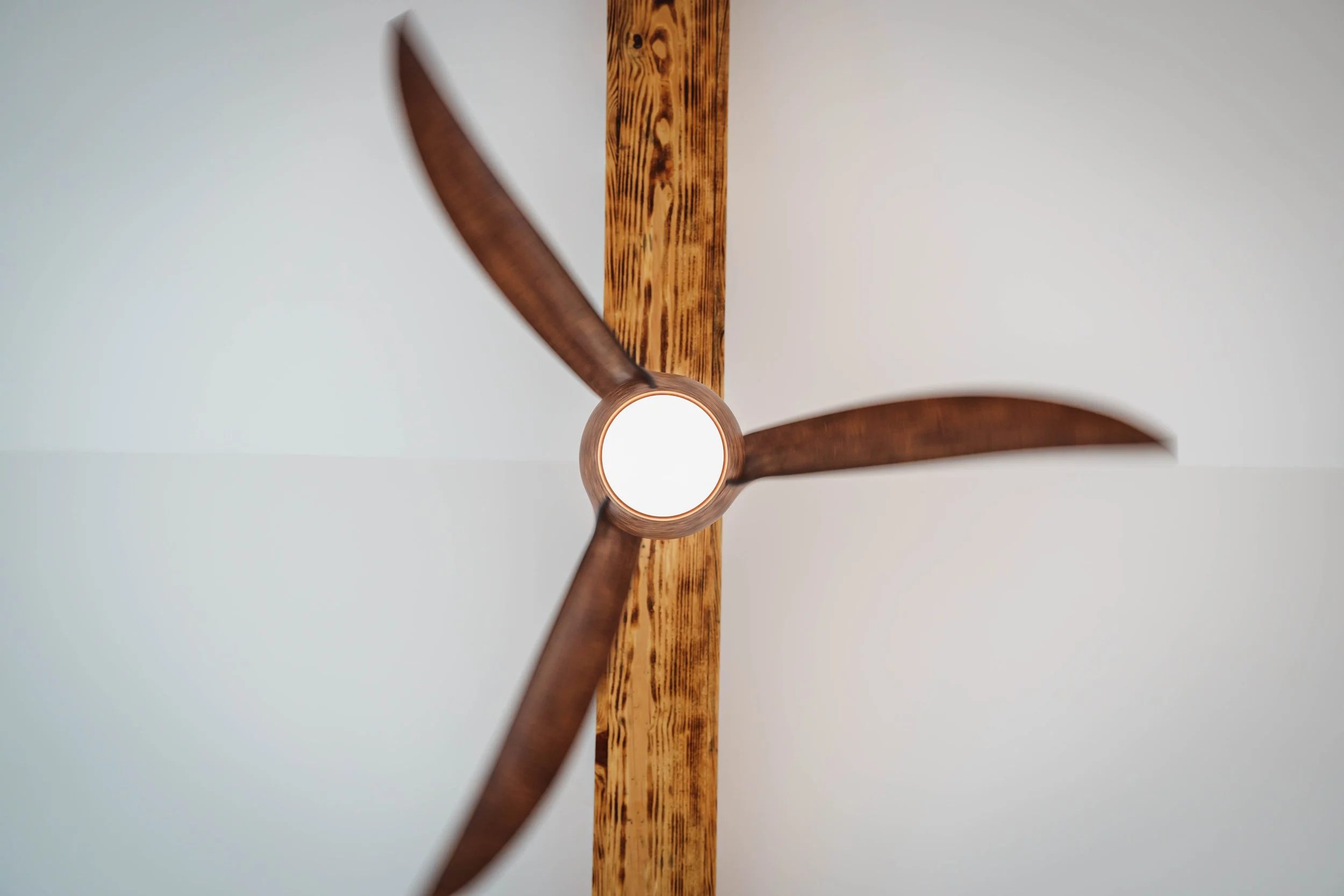 View of a ceiling fan with three wooden blades mounted on a wooden ceiling beam, seen from below.