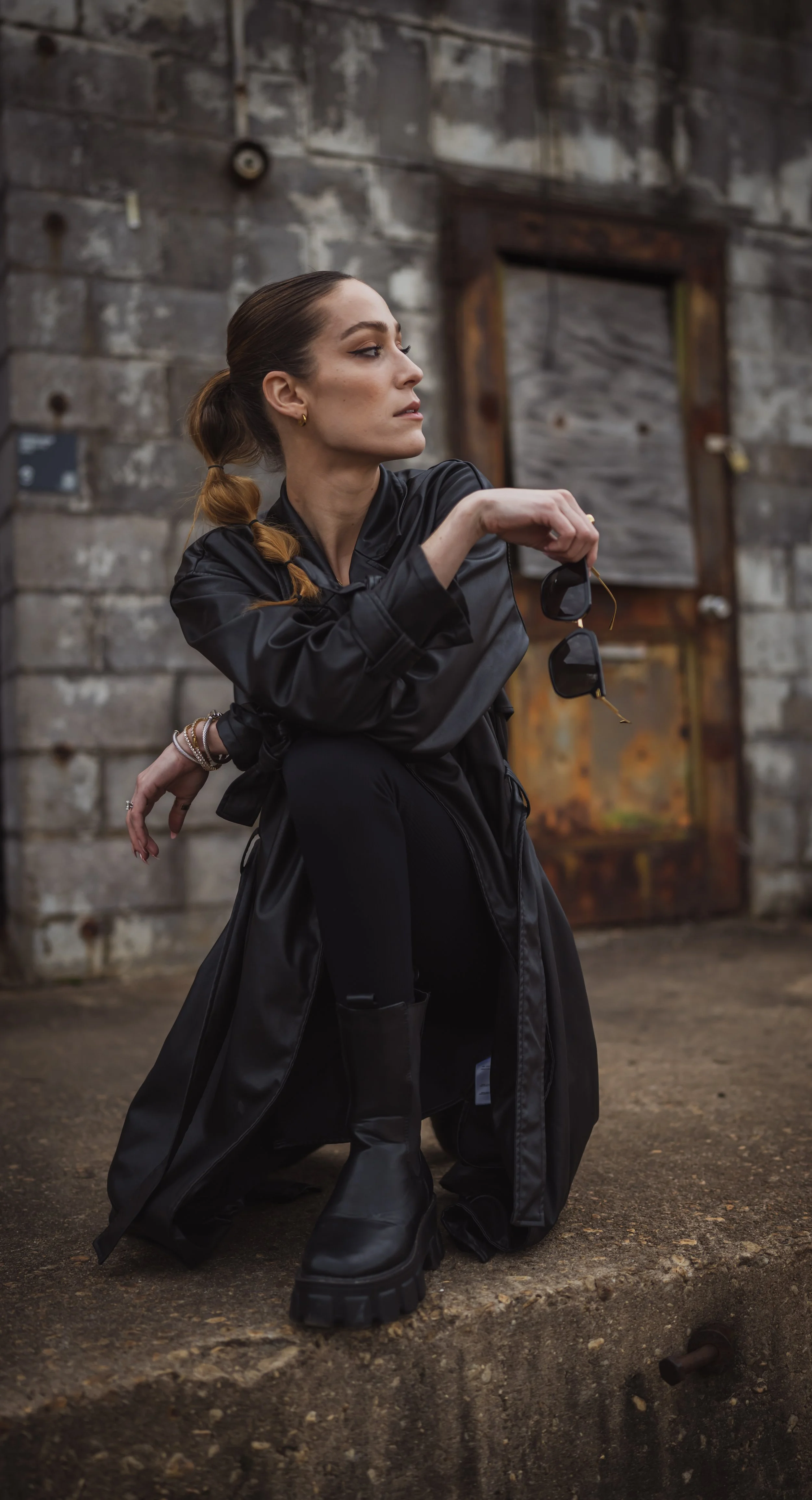 A woman with a ponytail, wearing a black leather jacket, black boots, and black pants, is crouching down outdoors on a gritty surface. She holds sunglasses in her left hand and appears to be looking to her left against a weathered, industrial brick b