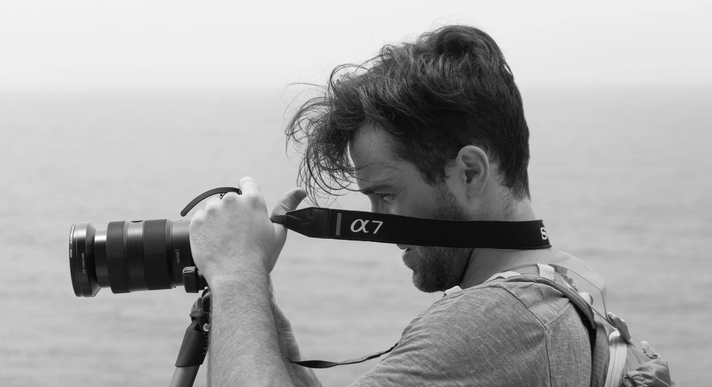 A man with disheveled hair looking through a camera lens mounted on a tripod, background of water or ocean, in black and white.
