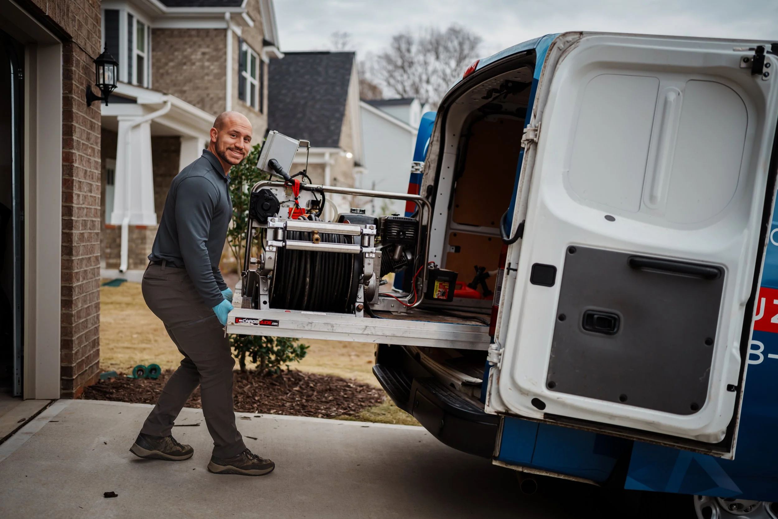 A man in work clothes and gloves is loading equipment into the back of a delivery van parked outside a house.