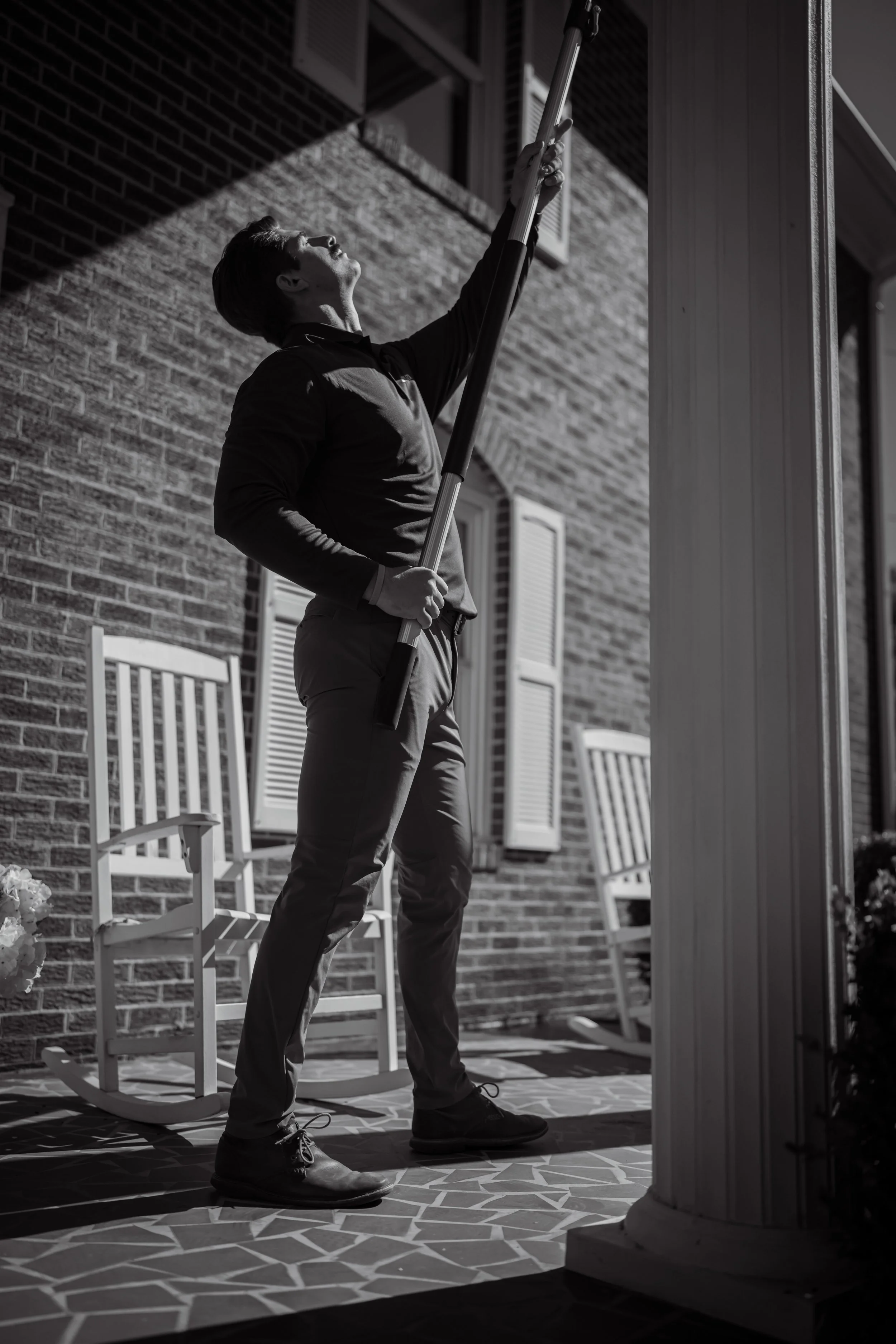 A man standing on a porch, reaching up with a pole to clean a window, with a brick wall and white rocking chairs in the background.