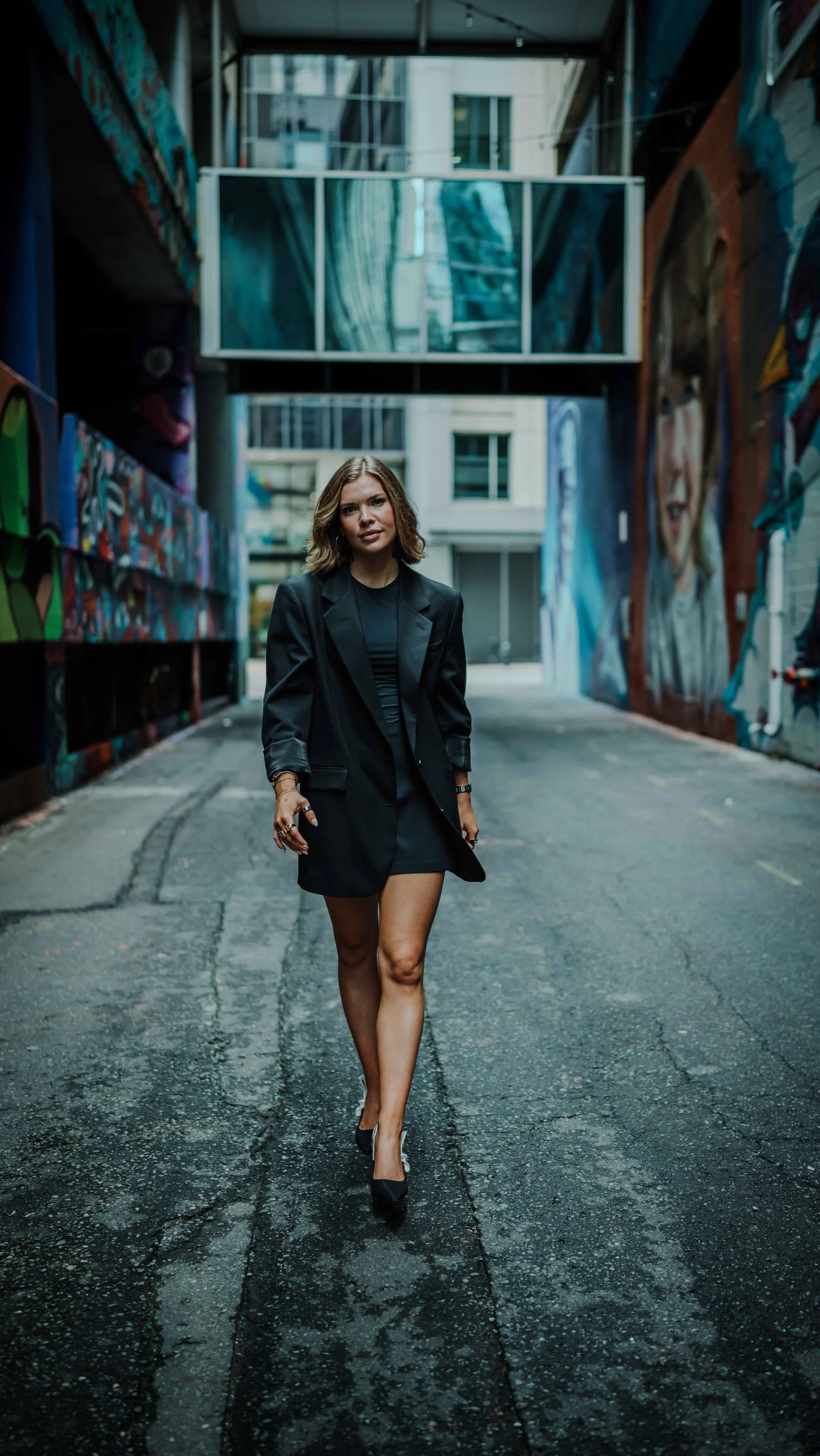 A young woman in a black blazer and high heels walking in an alleyway with colorful murals on the walls.