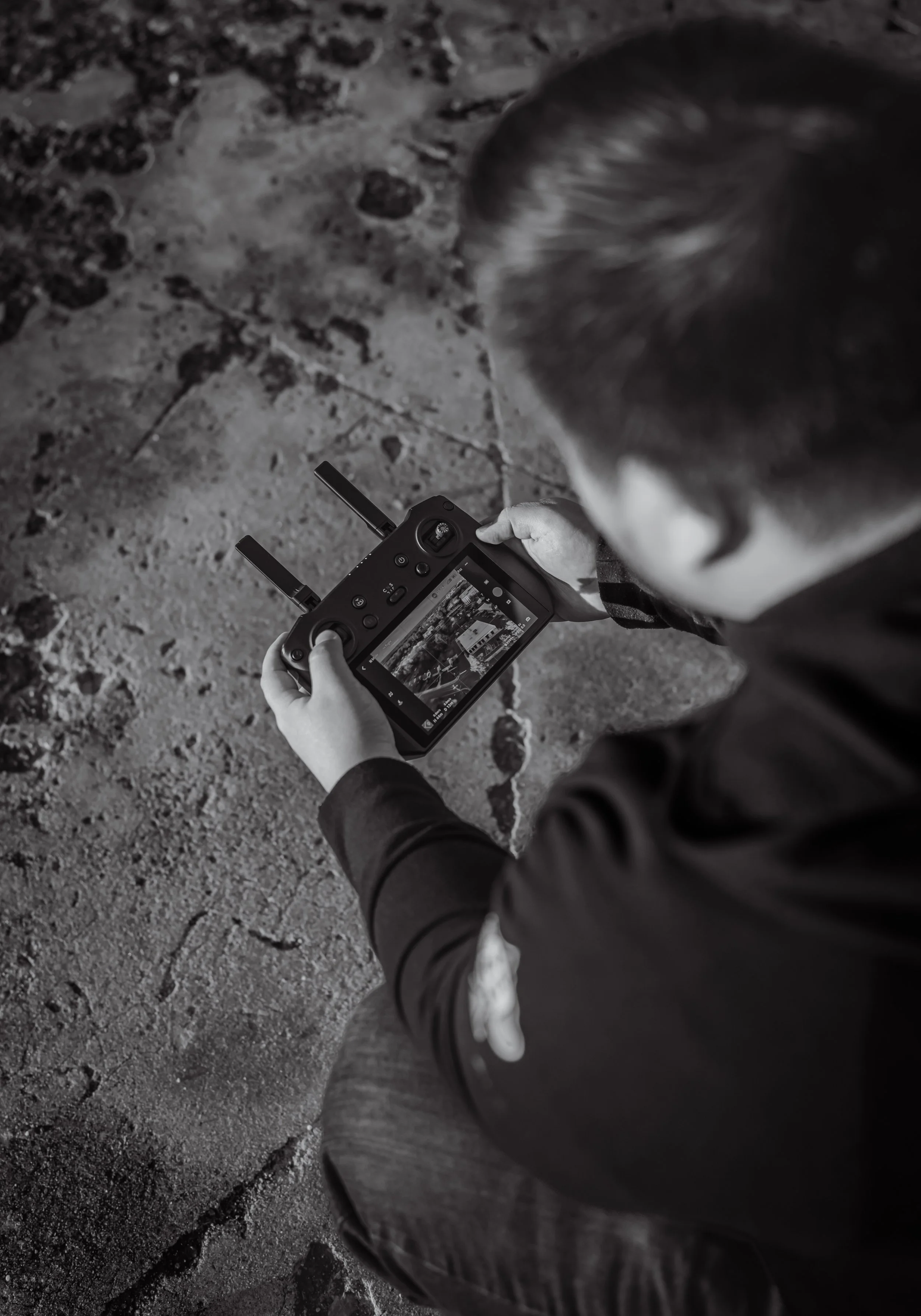 A young boy in a dark jacket holding a drone remote control, looking at a screen displaying a landscape photo, while sitting on a rough, rocky ground.