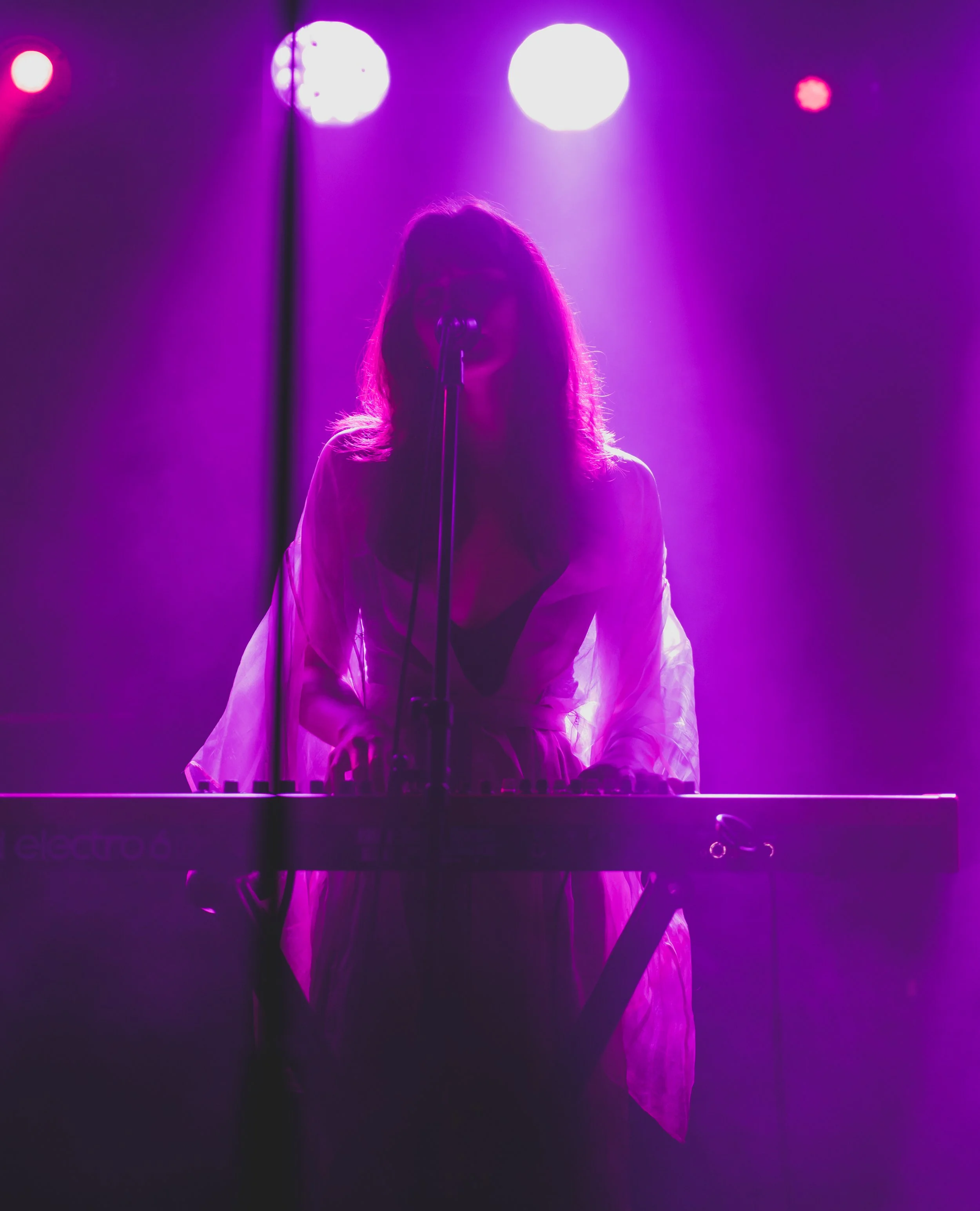 A woman performing on stage with purple lighting, playing a keyboard, and singing into a microphone.