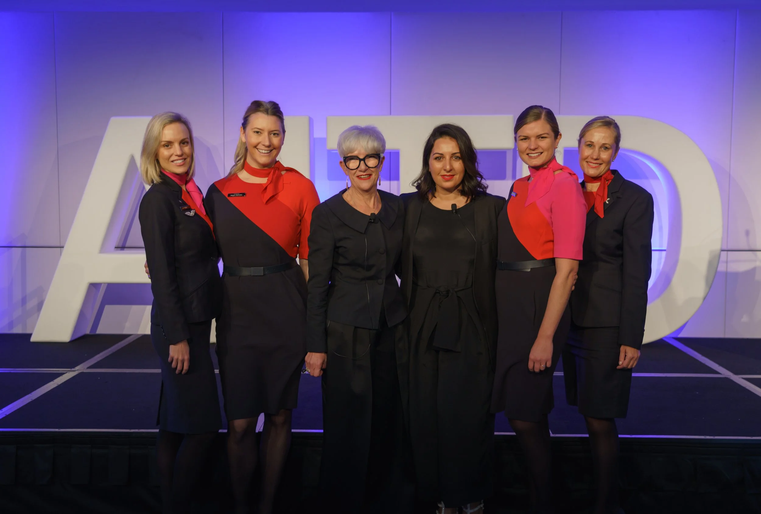 Six women standing in front of a large white letter 'AIDS' sign at an event, dressed in professional attire with some wearing black and others in red and pink blouses, smiling at the camera.