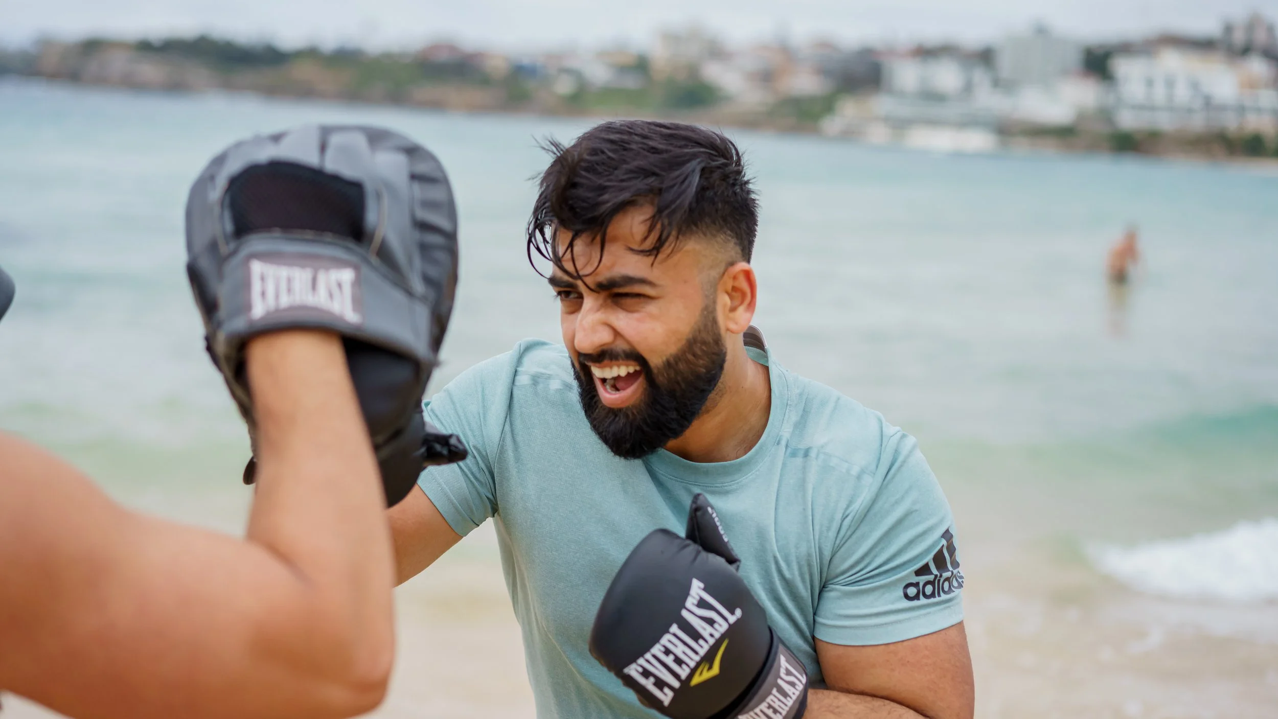 Two men engaging in boxing practice on the beach, one wearing boxing gloves and the other with boxing mitts, with a body of water and a distant shoreline in the background.