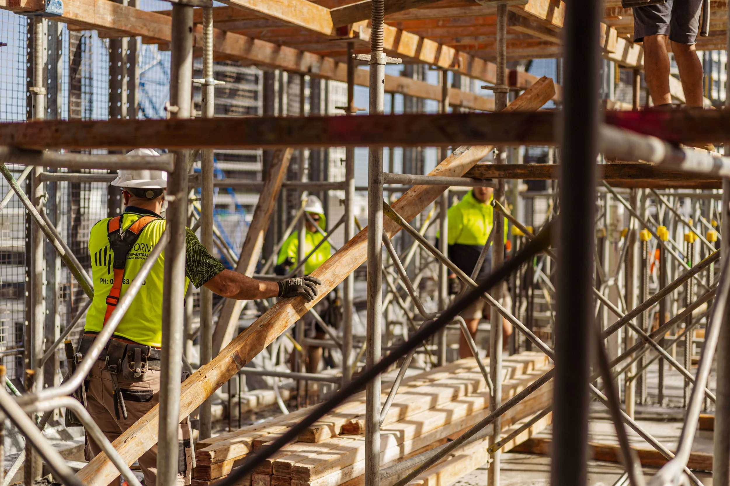 Construction workers assembling wooden scaffolding on a building site, surrounded by metal frames and safety gear.