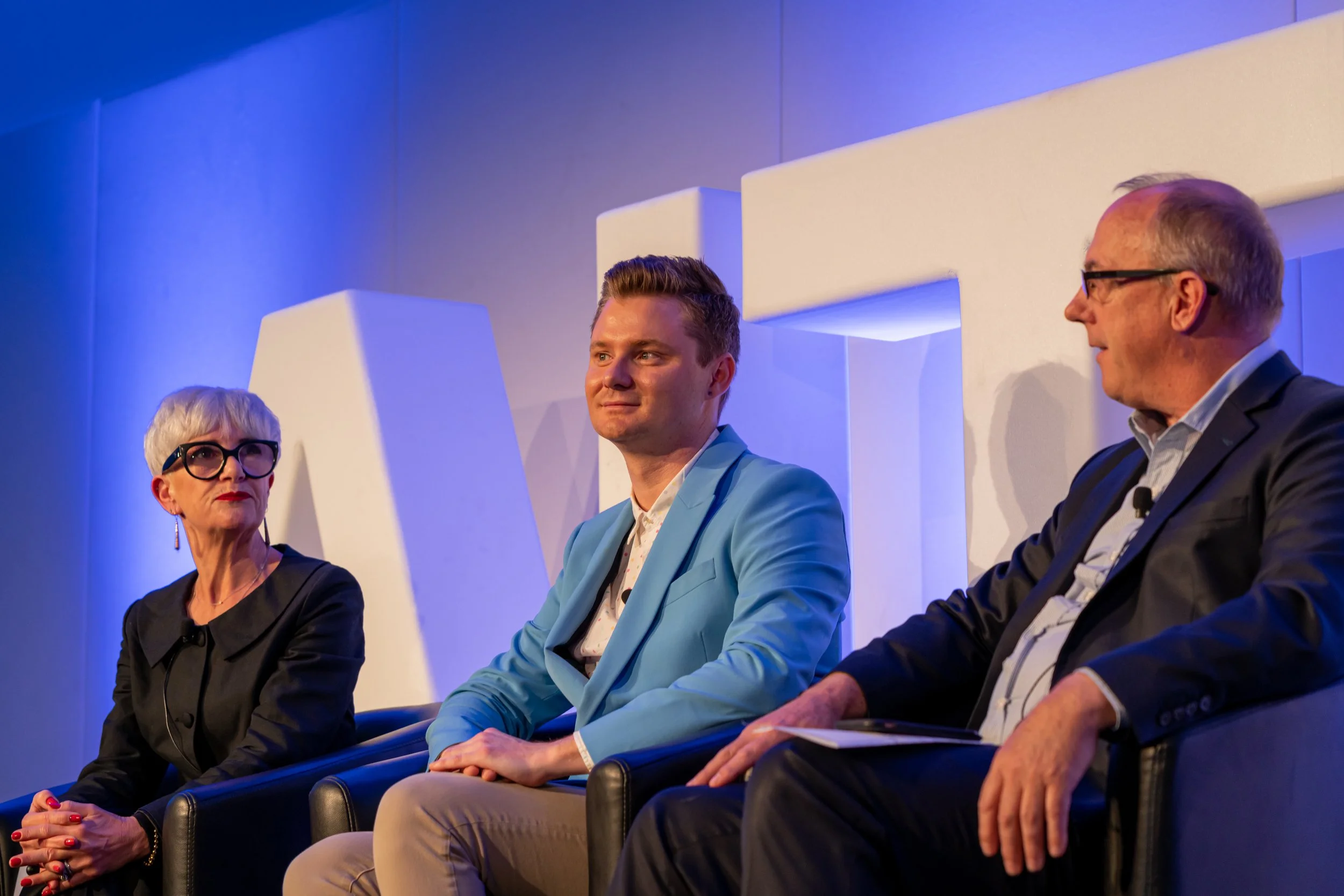 Three people seated on a stage during a panel discussion, with a woman on the left with short gray hair and glasses, a man in the middle with short brown hair in a blue blazer, and an older man on the right with glasses in a dark suit, all engaged in