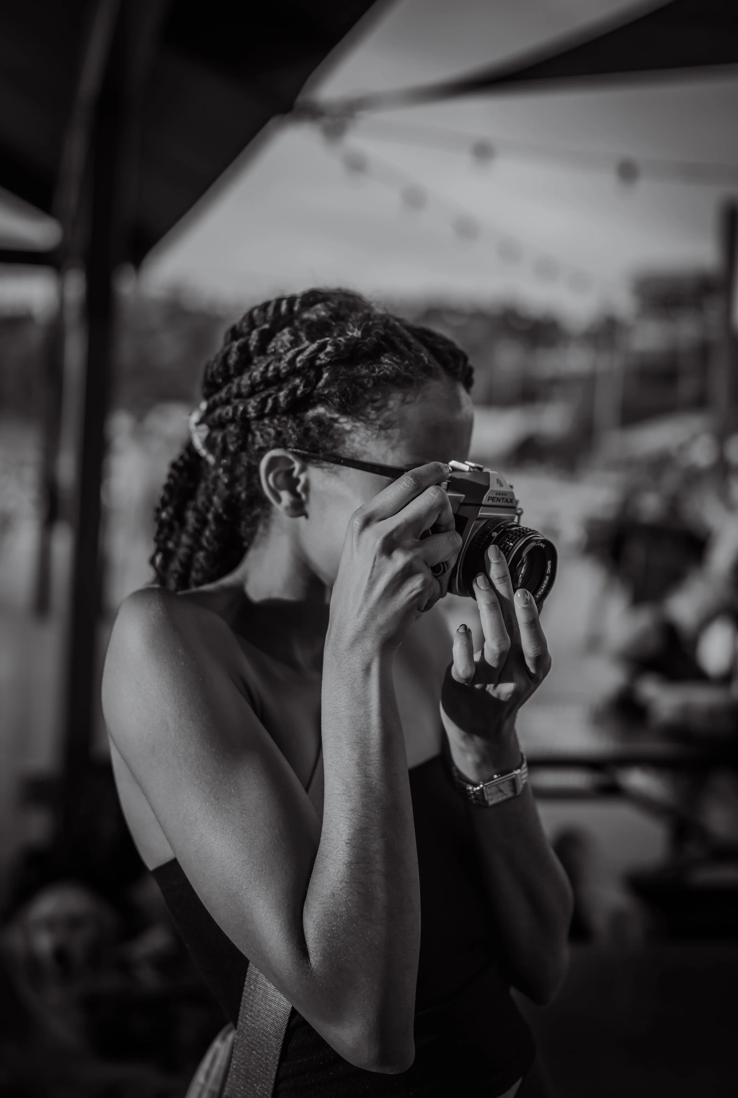 A woman with braided hair and glasses taking a black and white photo with a vintage film camera outdoors.