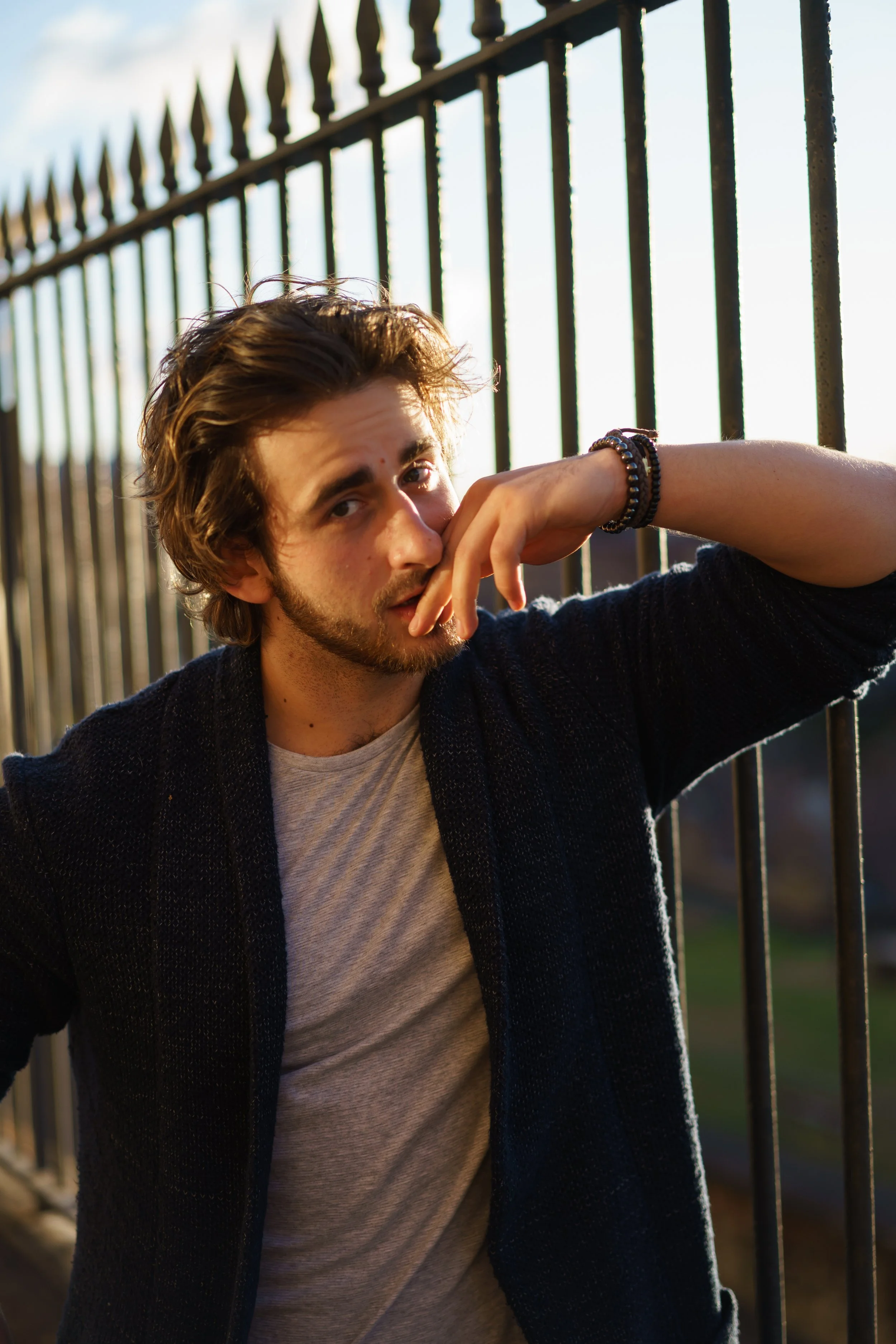 A young man with brown hair and a beard leaning against a metal fence during sunset, wearing a beige shirt and a dark cardigan, with his hand near his face.