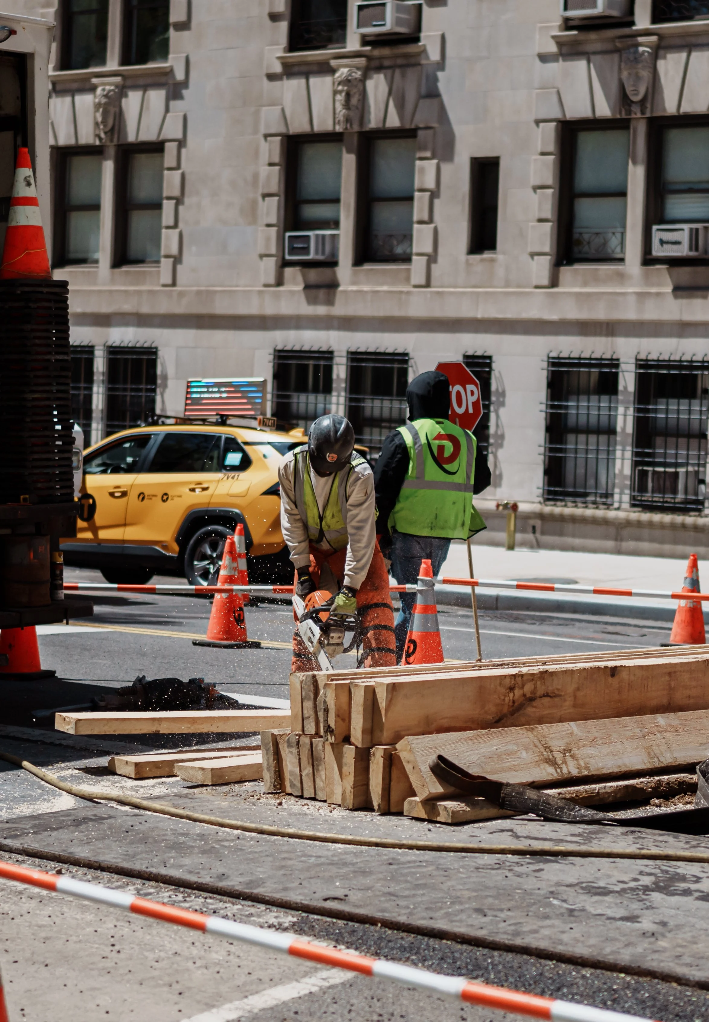 Construction workers cutting wood on a city street with traffic cones and a yellow taxi in the background.