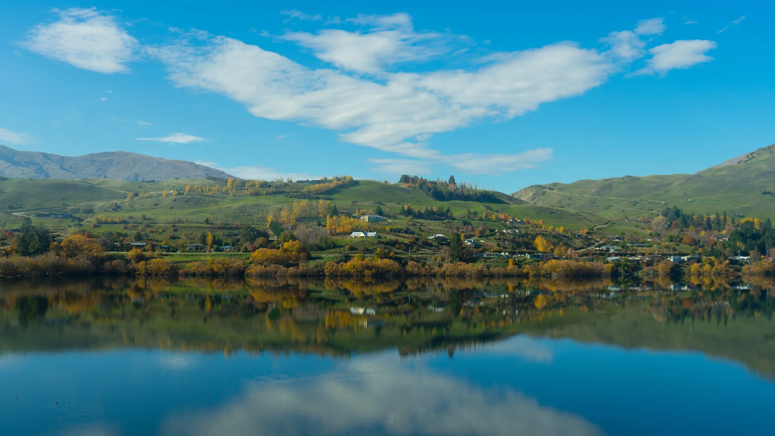 Scenic view of a calm lake reflecting green hills, trees with fall foliage, and a partly cloudy blue sky.
