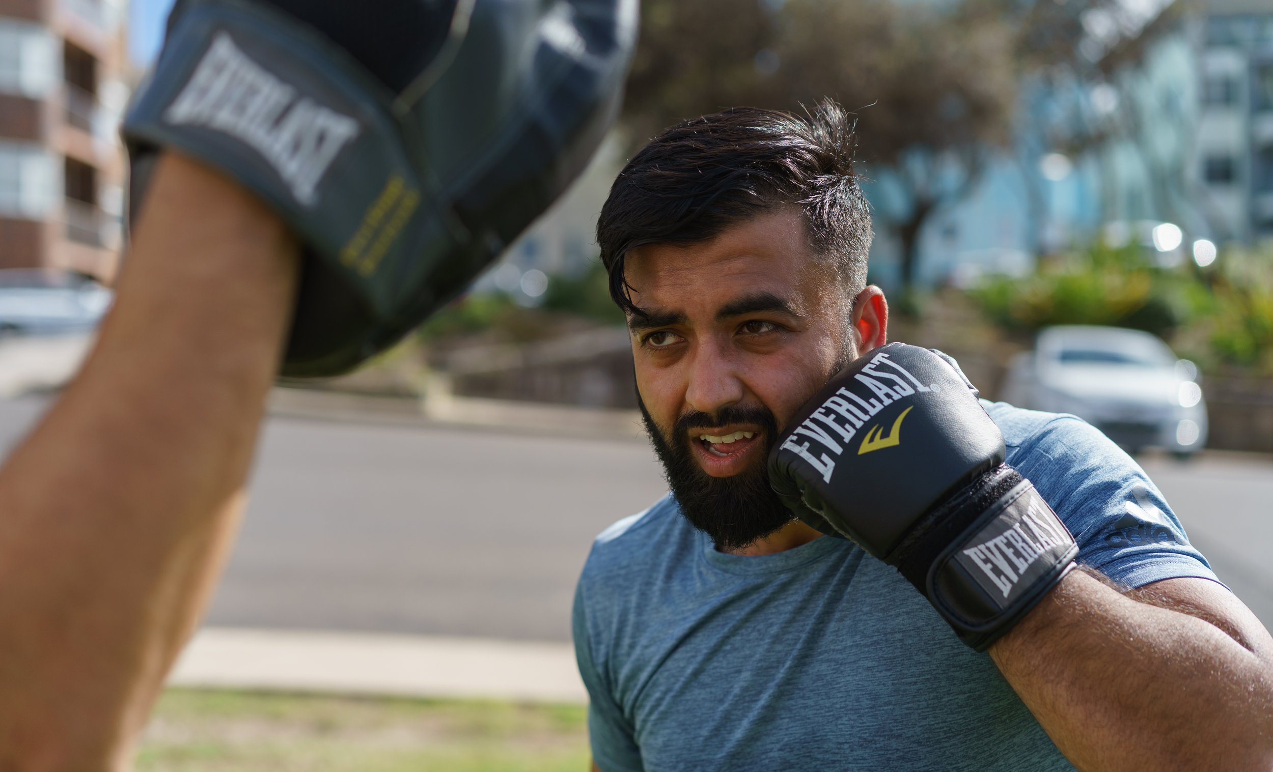 A man doing boxing training outdoors, wearing black Everlast boxing gloves and a blue shirt.