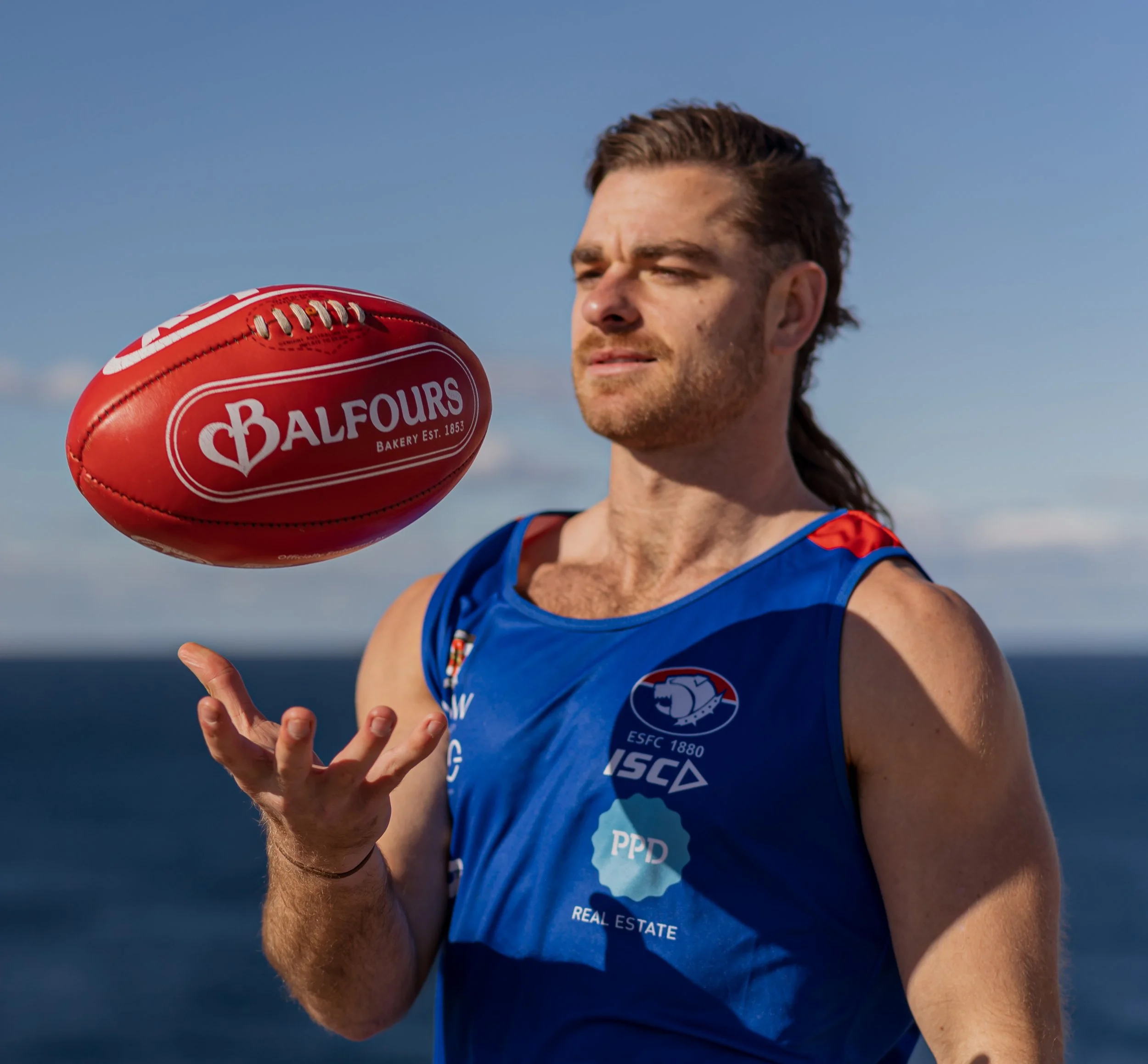Male athlete spinning a red football on his finger outdoors, wearing a blue sports jersey, with sky and sea in background.