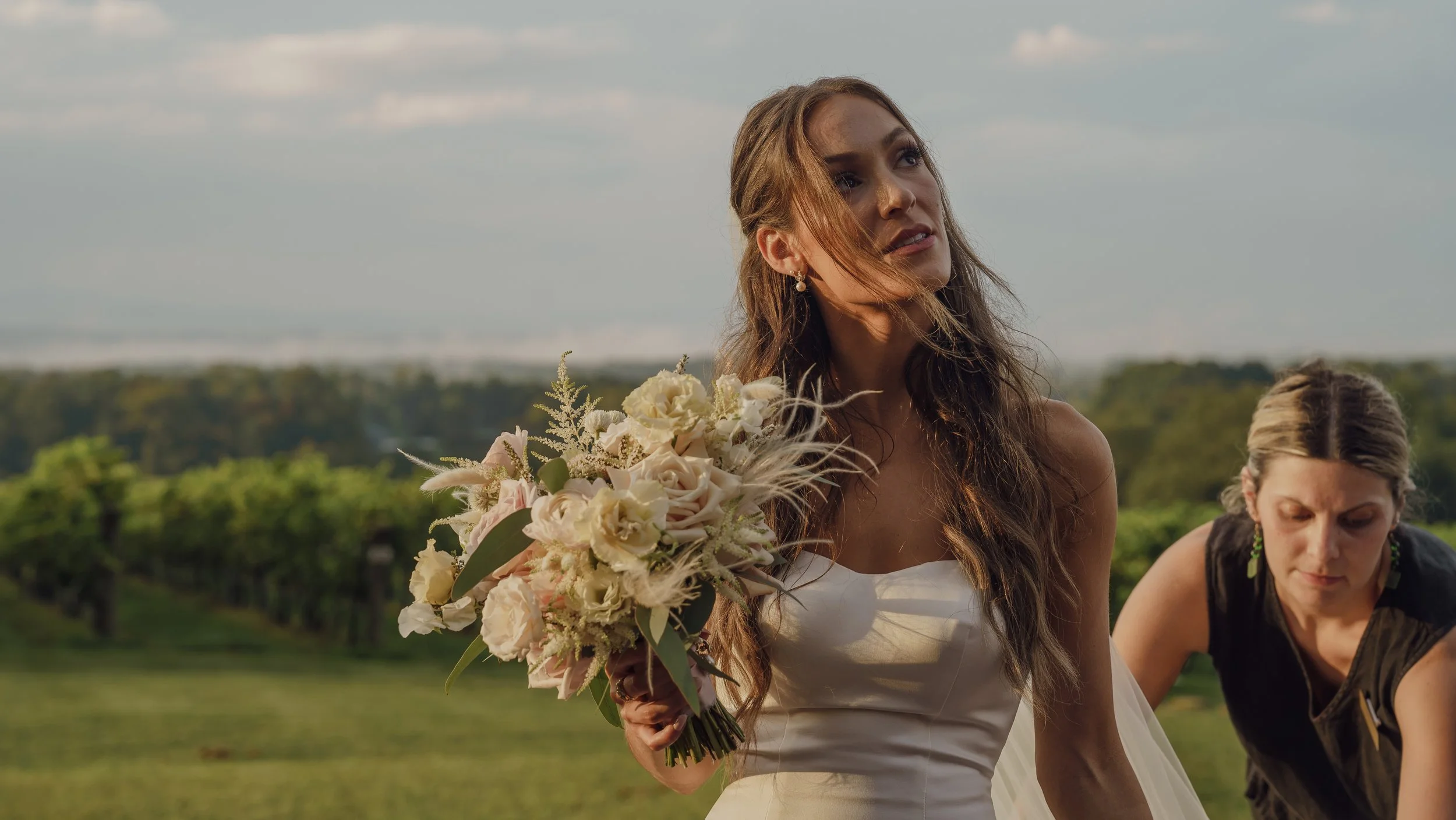 A woman in a wedding dress holding a bouquet of white and cream flowers during an outdoor setting with green fields and trees in the background.
