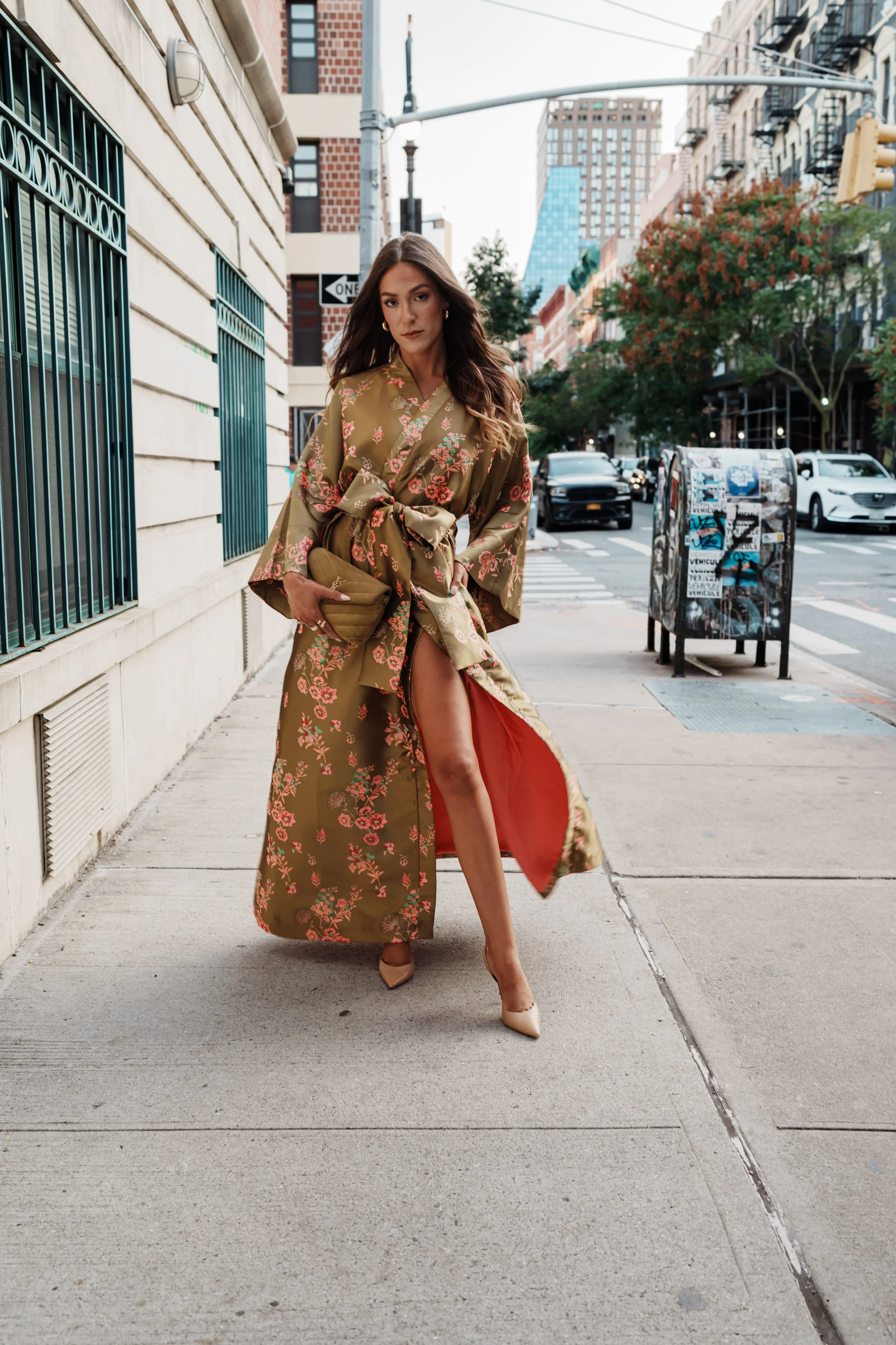 A woman in a floral kimono-style dress with a high slit walking on the sidewalk in an urban street.