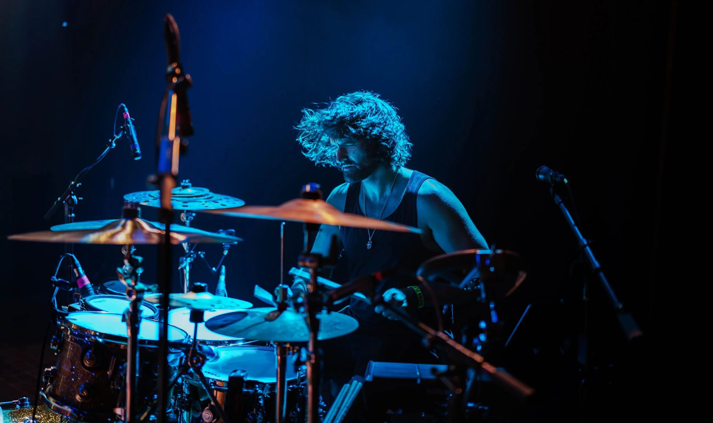 A drummer performing on stage under blue lighting, surrounded by drums and percussion instruments, with dark background.