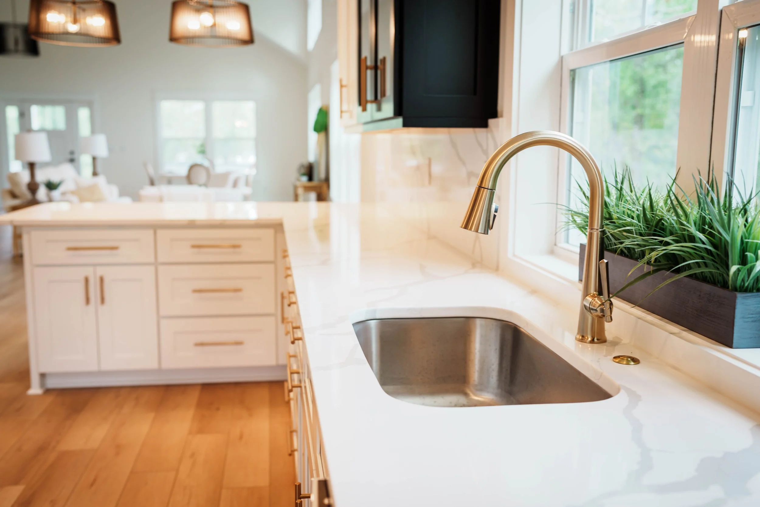 Modern kitchen with white marble countertop, brass faucet, stainless steel sink, and decor with green plants. Background shows a spacious living area with windows and furniture.