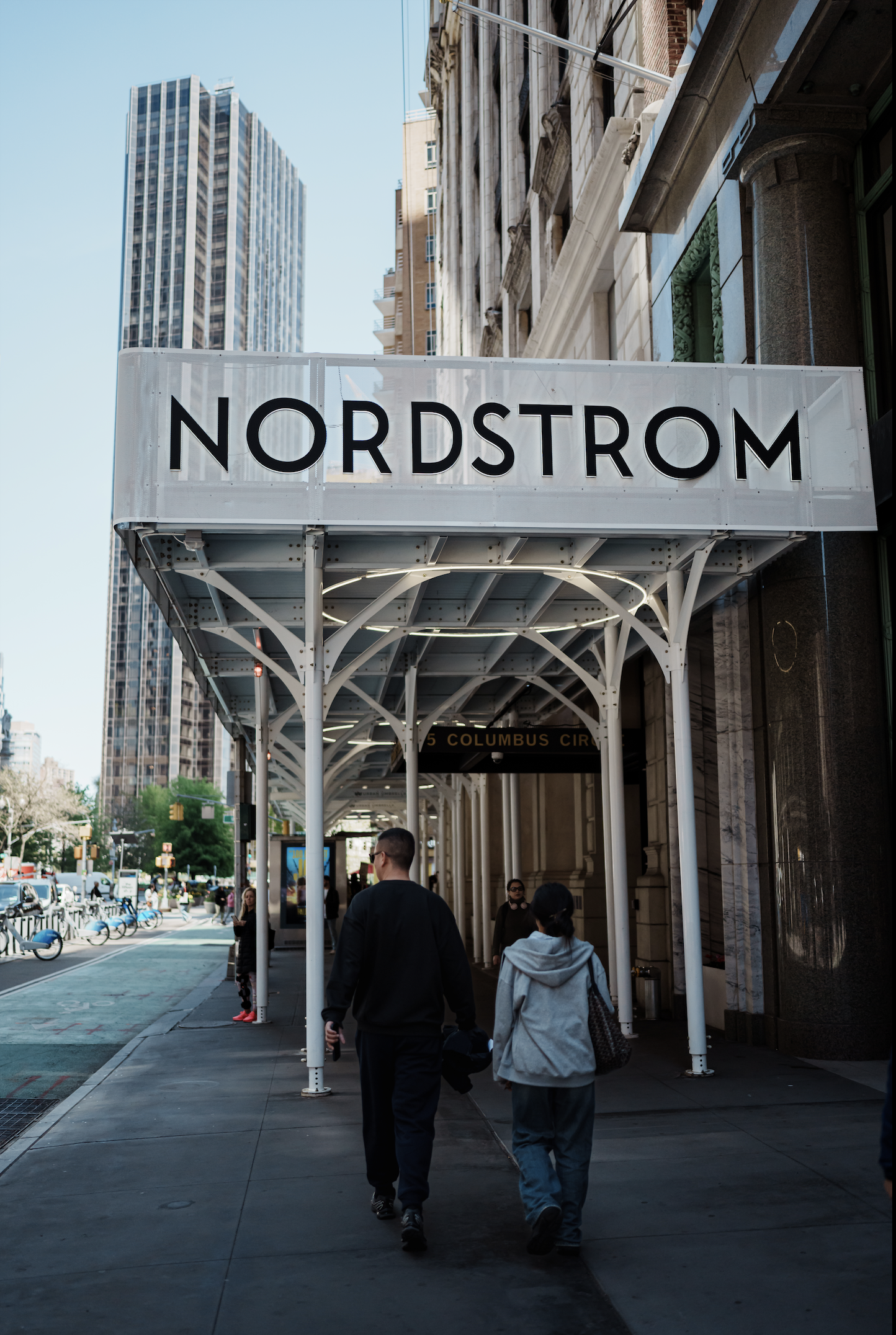 People walking past the Nordstrom store entrance on a city street with tall buildings in the background.