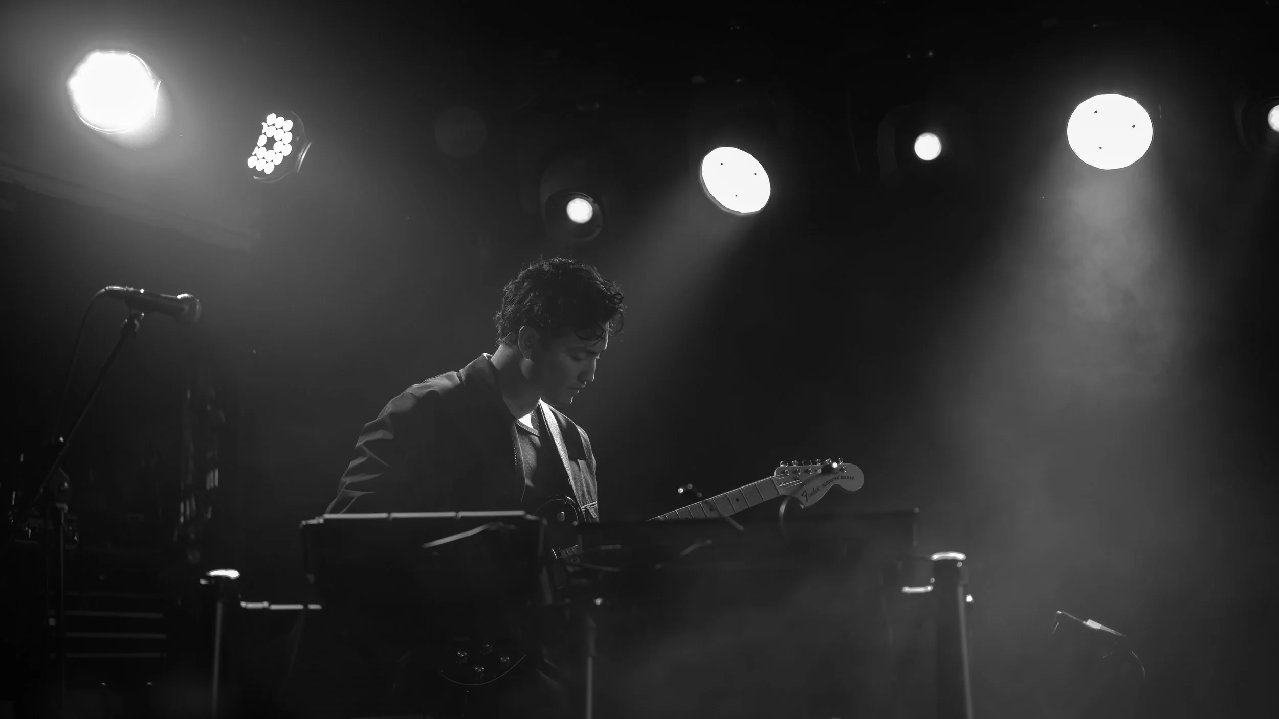 A black-and-white photo of a young man playing an electric guitar on stage, illuminated by several stage lights above him, with a microphone stand nearby.